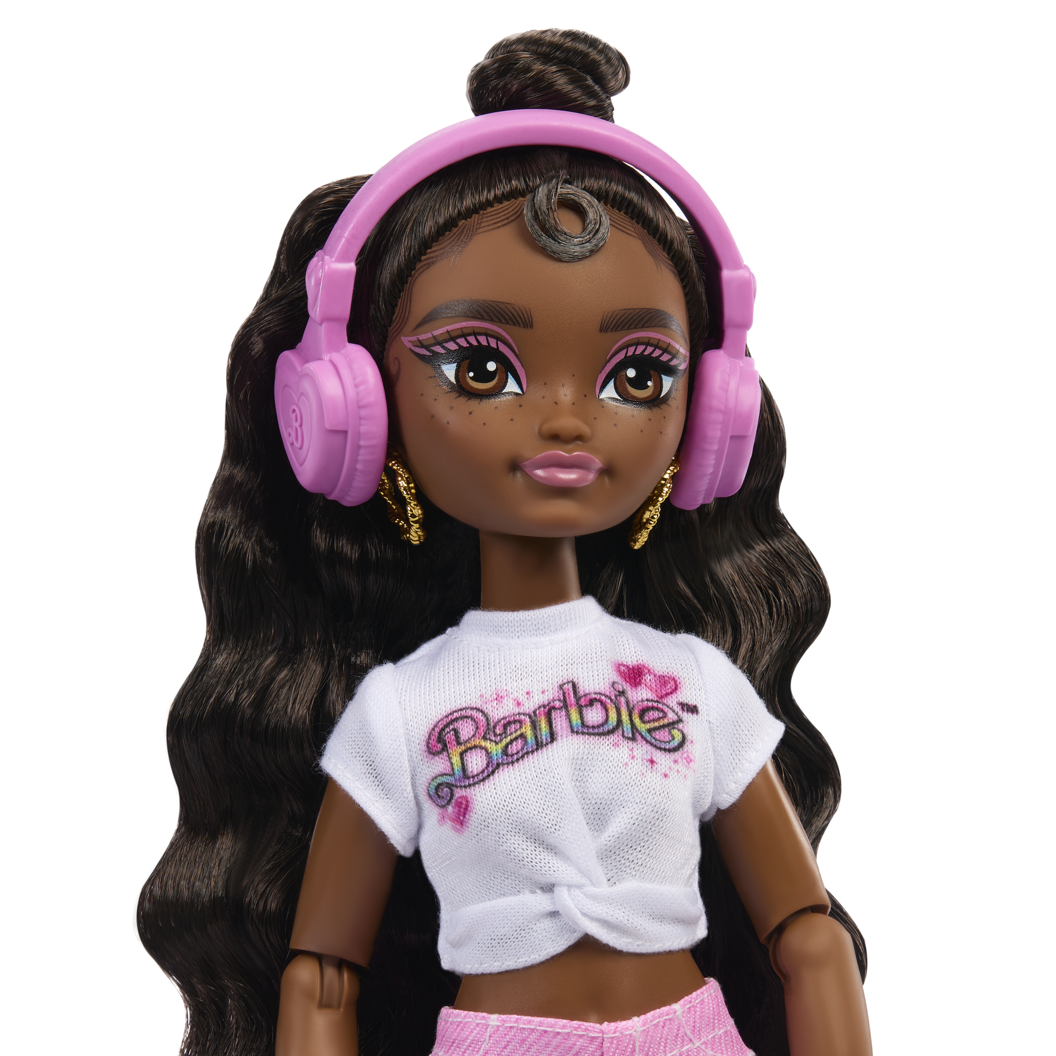 Una muñeca Barbie de piel oscura, con camisa blanca, pantalones cortos rosas y auriculares rosas. Pelo largo, oscuro y ondulado.