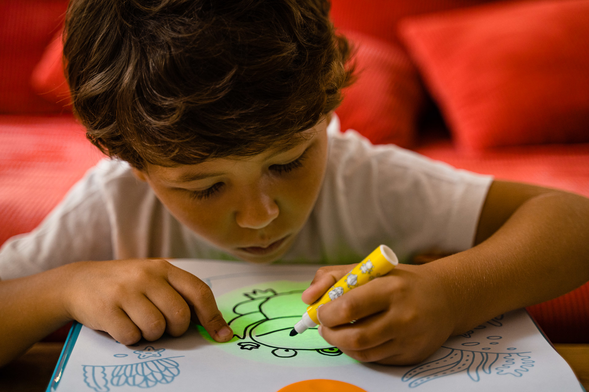 Dziecko rysuje żółtym markerem. Rysunek to zielona postać z kreskówki. Tło to czerwona sofa.
