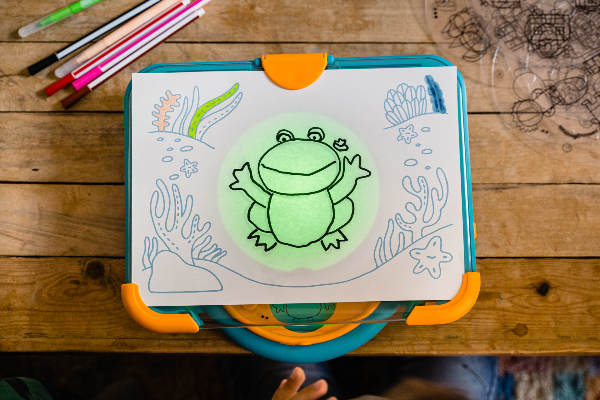 Tablica do rysowania z ilustracją żaby i markerami na drewnianej powierzchni. Żaba świeci na zielono.