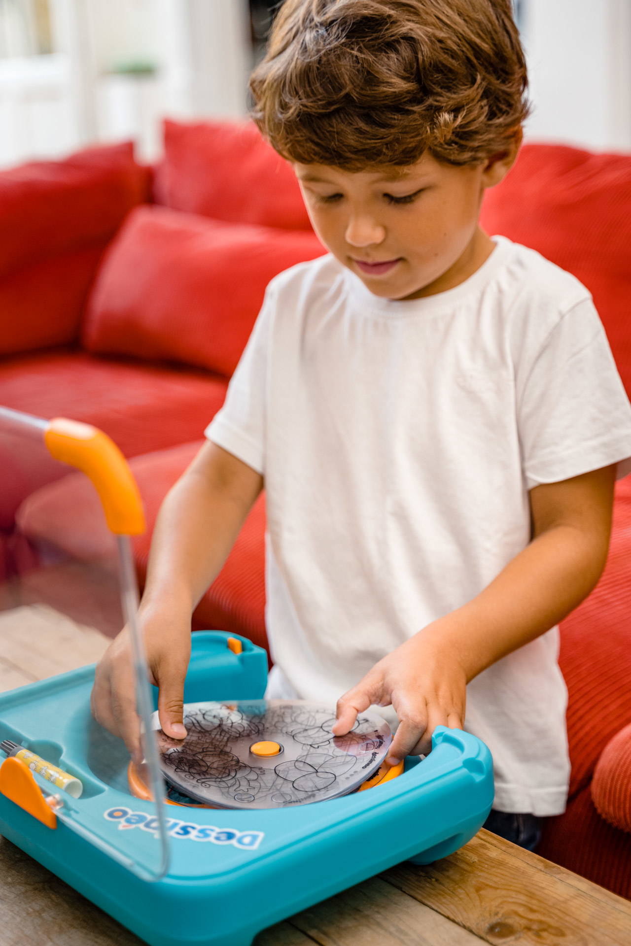 Chłopiec w białej koszulce używa zabawki do rysowania. Siedzi przed czerwoną sofą.