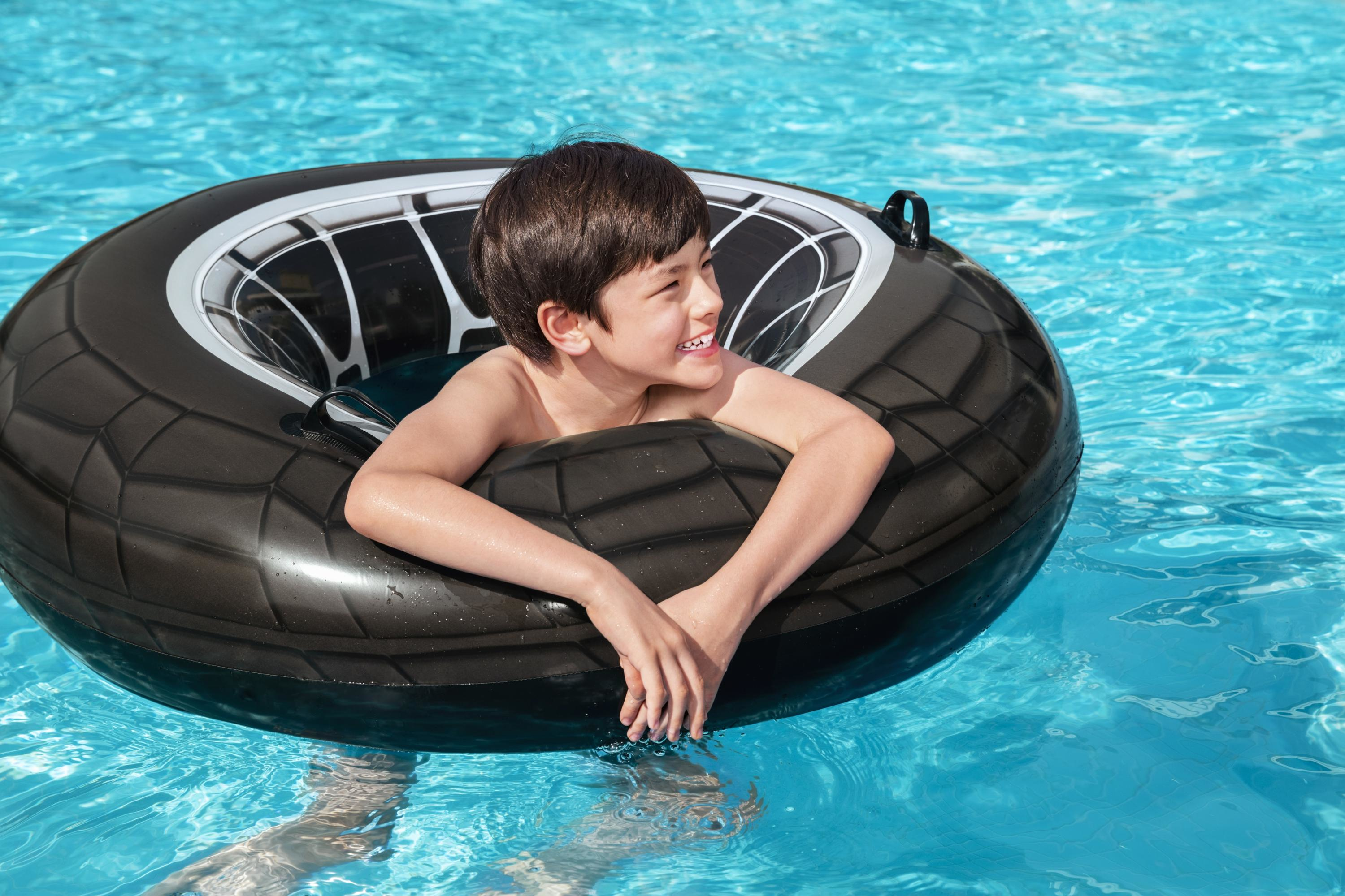 A boy smiles while floating in a pool on a black tire tube. The pool water is bright blue.