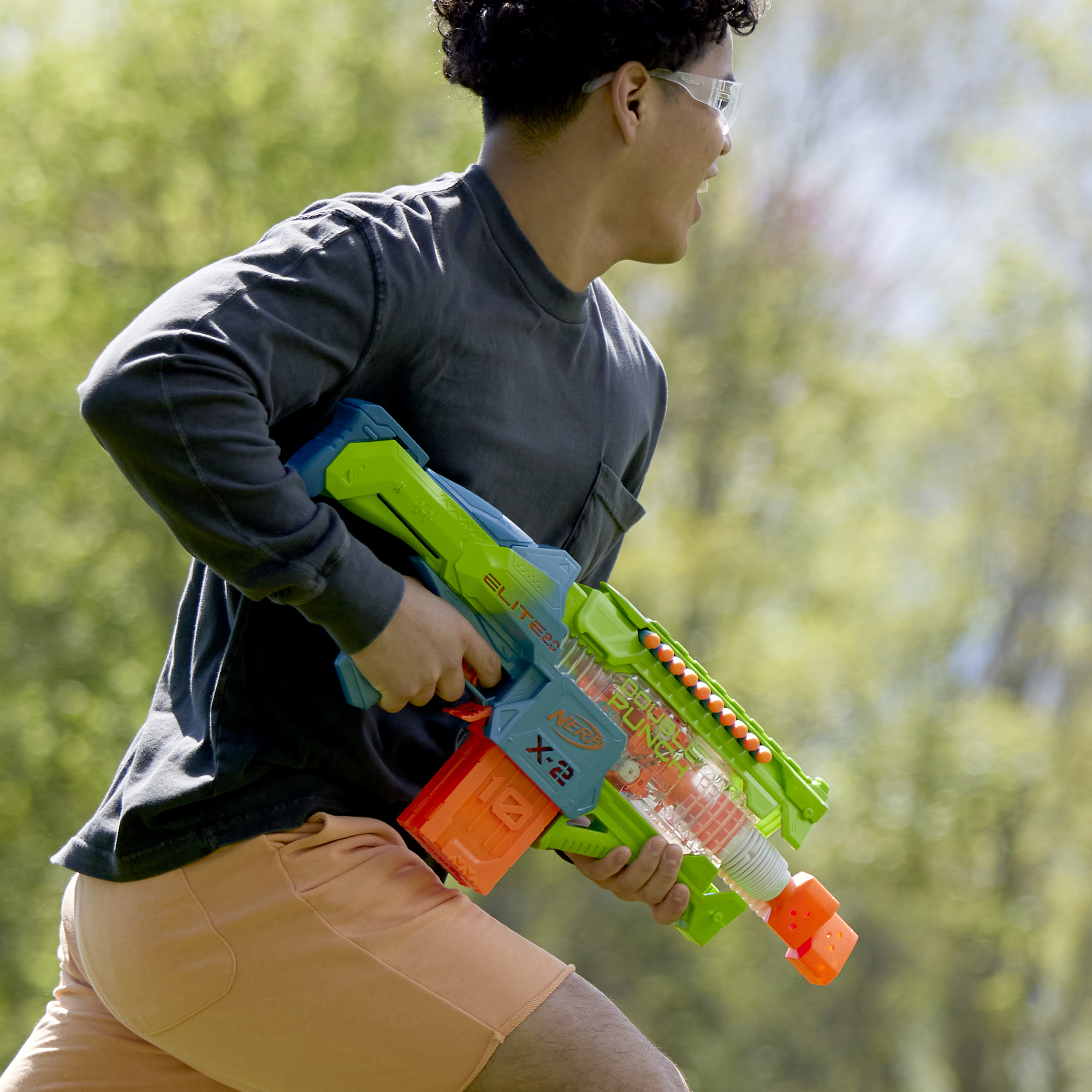 Una persona corre con una pistola giocattolo verde e blu. Indossa occhiali, una camicia nera e pantaloncini arancioni.
