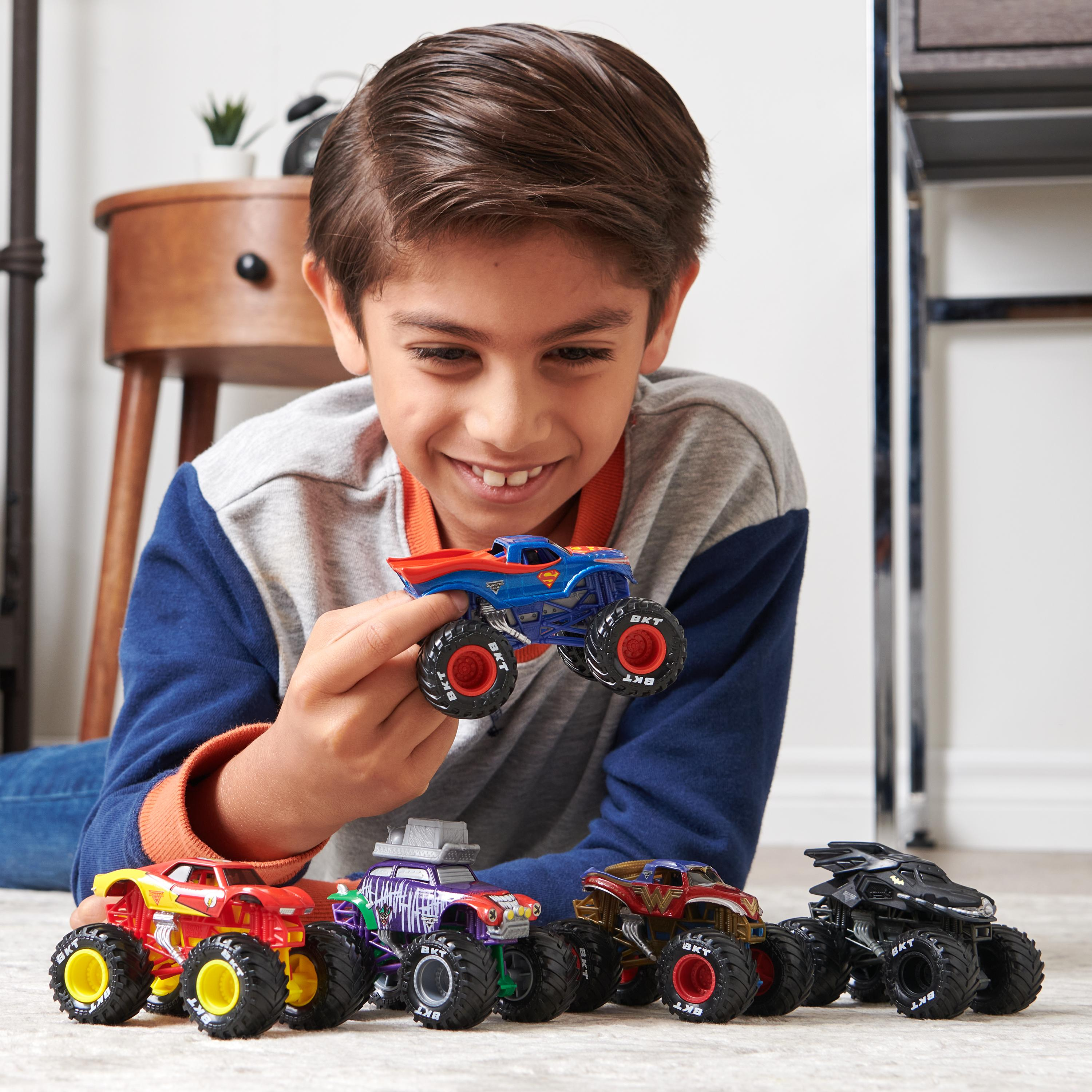 Ein Junge lächelt und hält einen Superman-Monstertruck. Andere Trucks stehen auf dem Boden; ein Nachttisch und der Boden sind im Hintergrund.