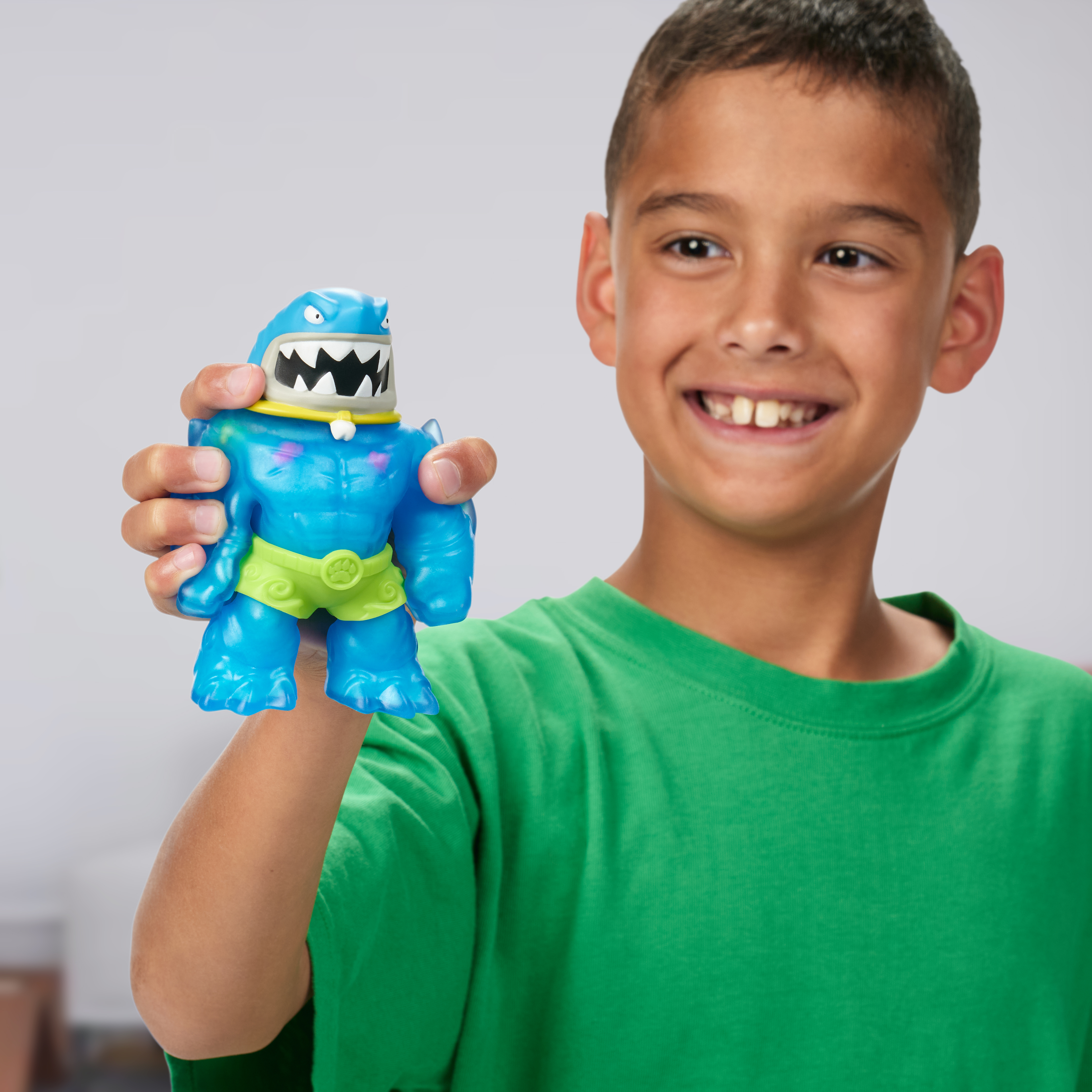 Ein Junge hält ein blaues Monstertoy. Der Junge lächelt, trägt ein grünes Hemd, vor grauem Hintergrund.