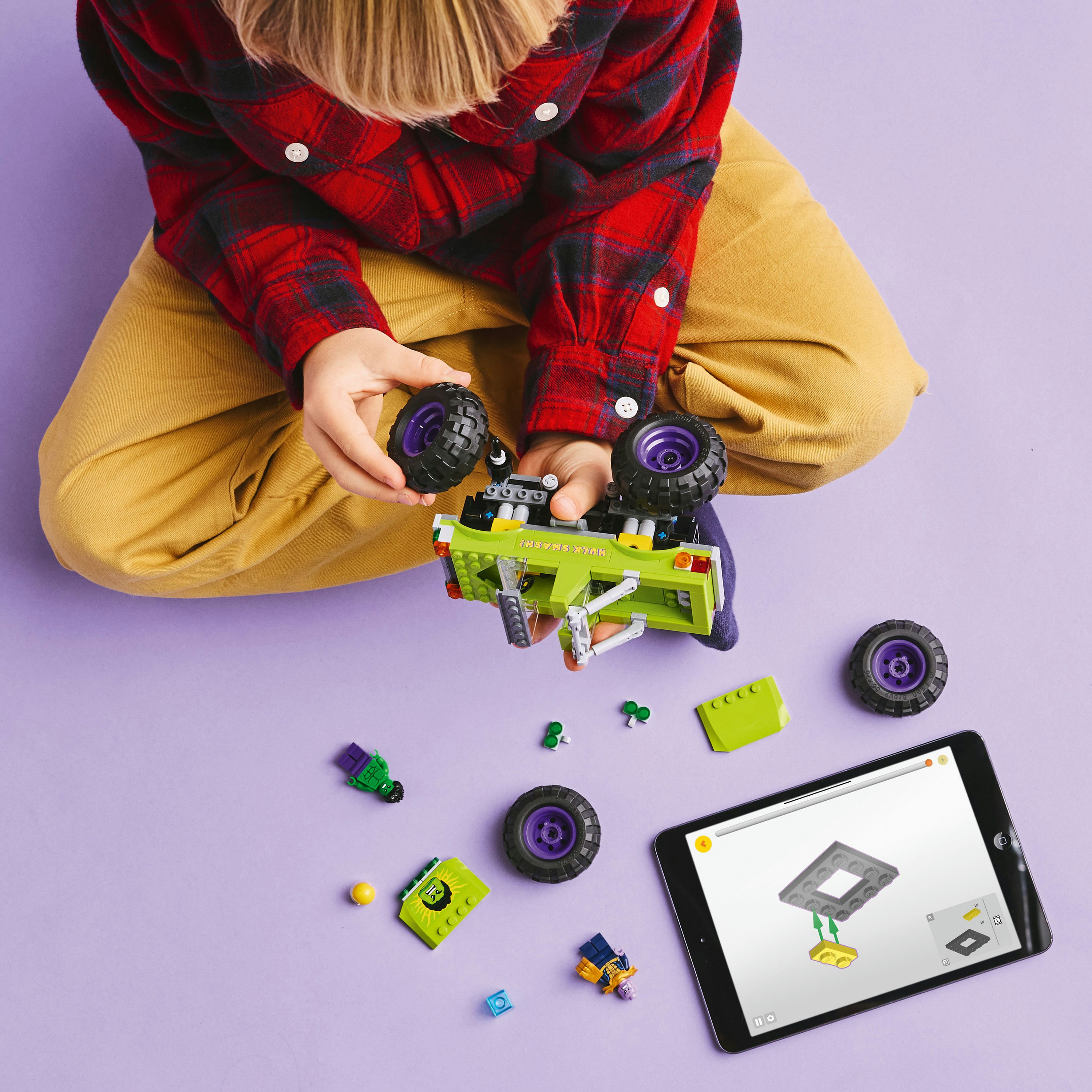 Dziecko w koszuli w kratę i beżowych spodniach buduje samochód Lego. Części i tablet z instrukcjami na fioletowej powierzchni.