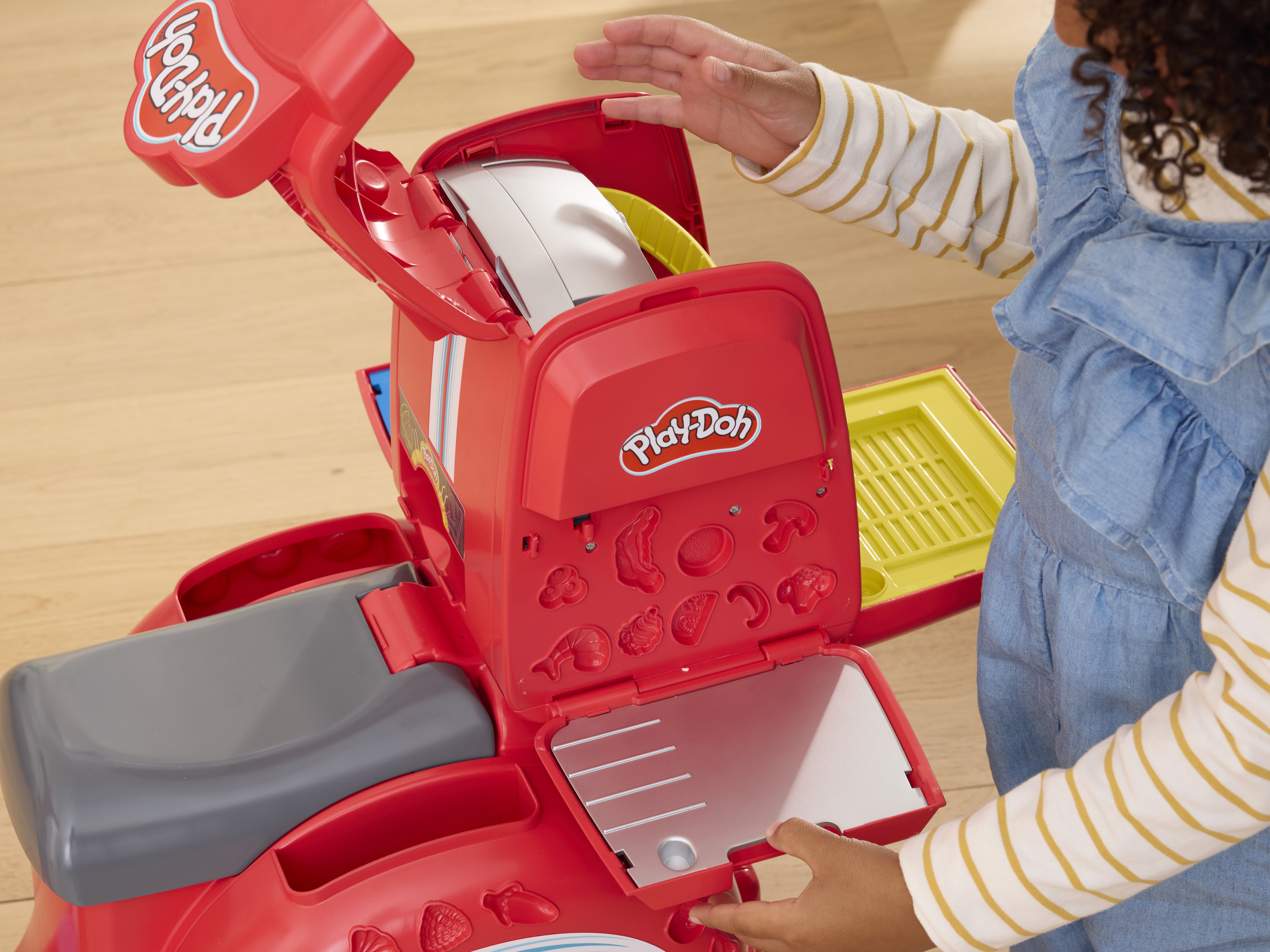 Un niño abre un juego de Play-Doh rojo. El juego tiene compartimentos y moldes para plastilina. Suelo de madera al fondo.