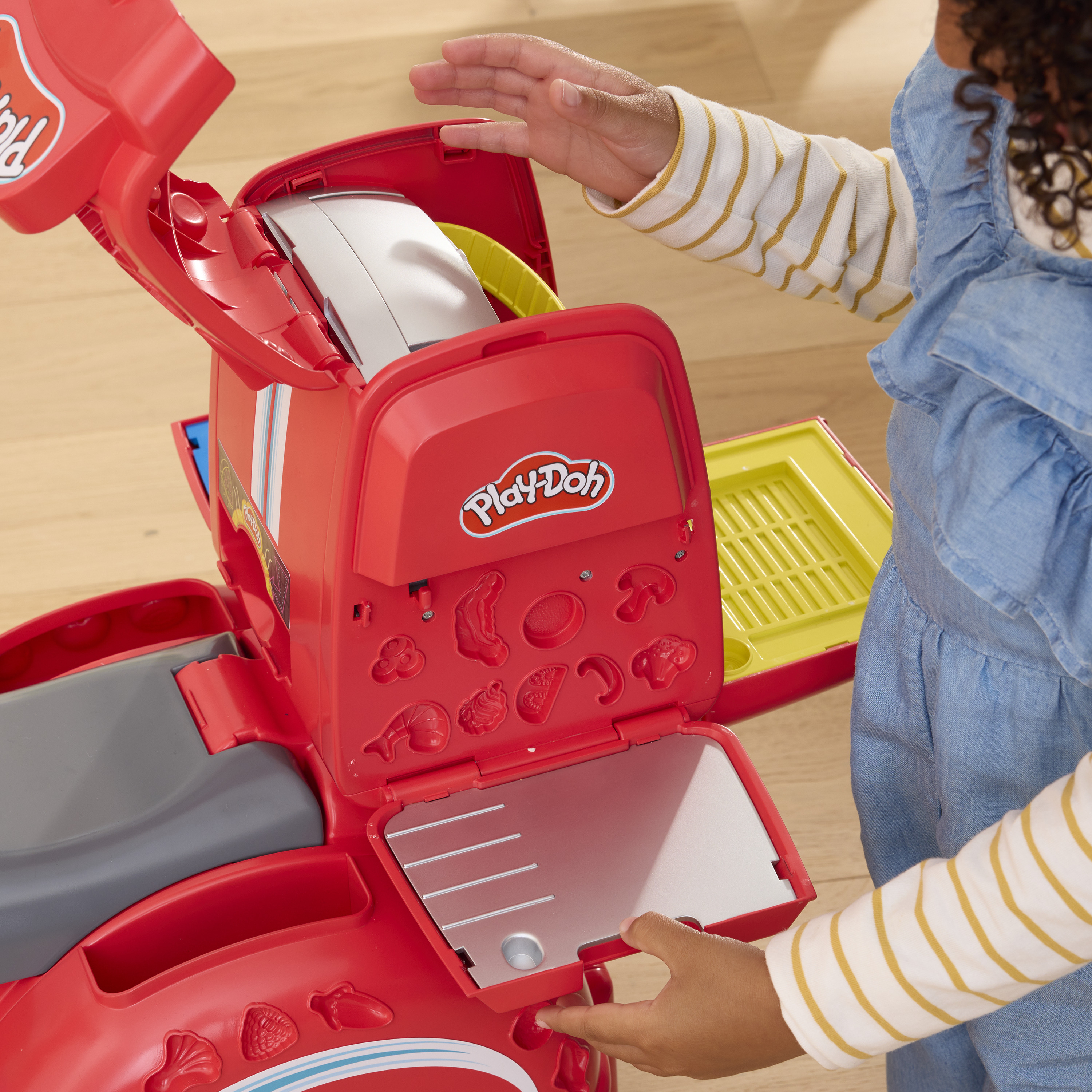 Un niño abre un juego de juguetes Play-Doh rojo. El juego tiene moldes de comida y compartimentos. El fondo es de madera.