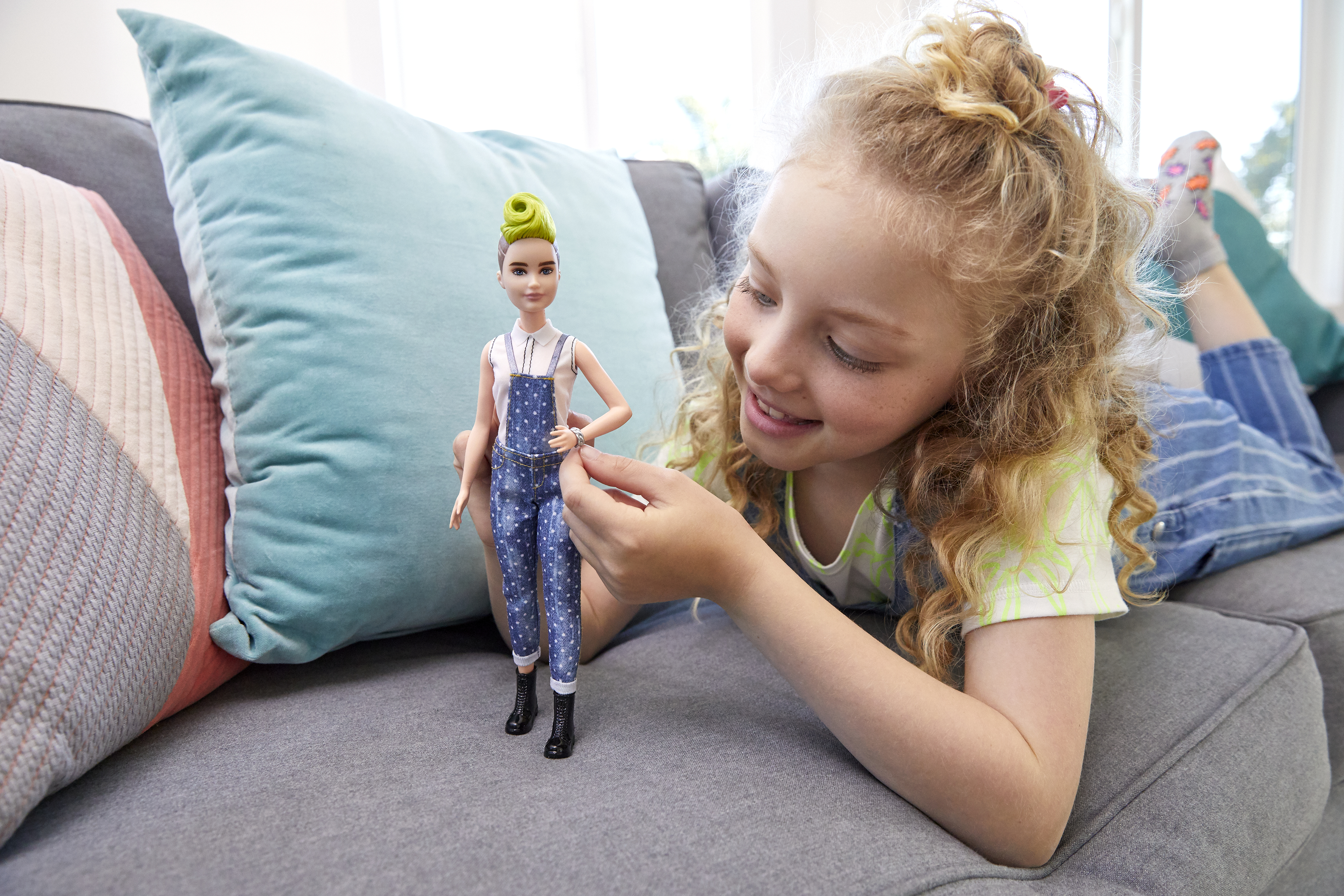 Ein Mädchen mit lockigem Haar betrachtet eine Puppe in blauen Overalls. Die Puppe liegt auf einem grauen Sofa mit Kissen.