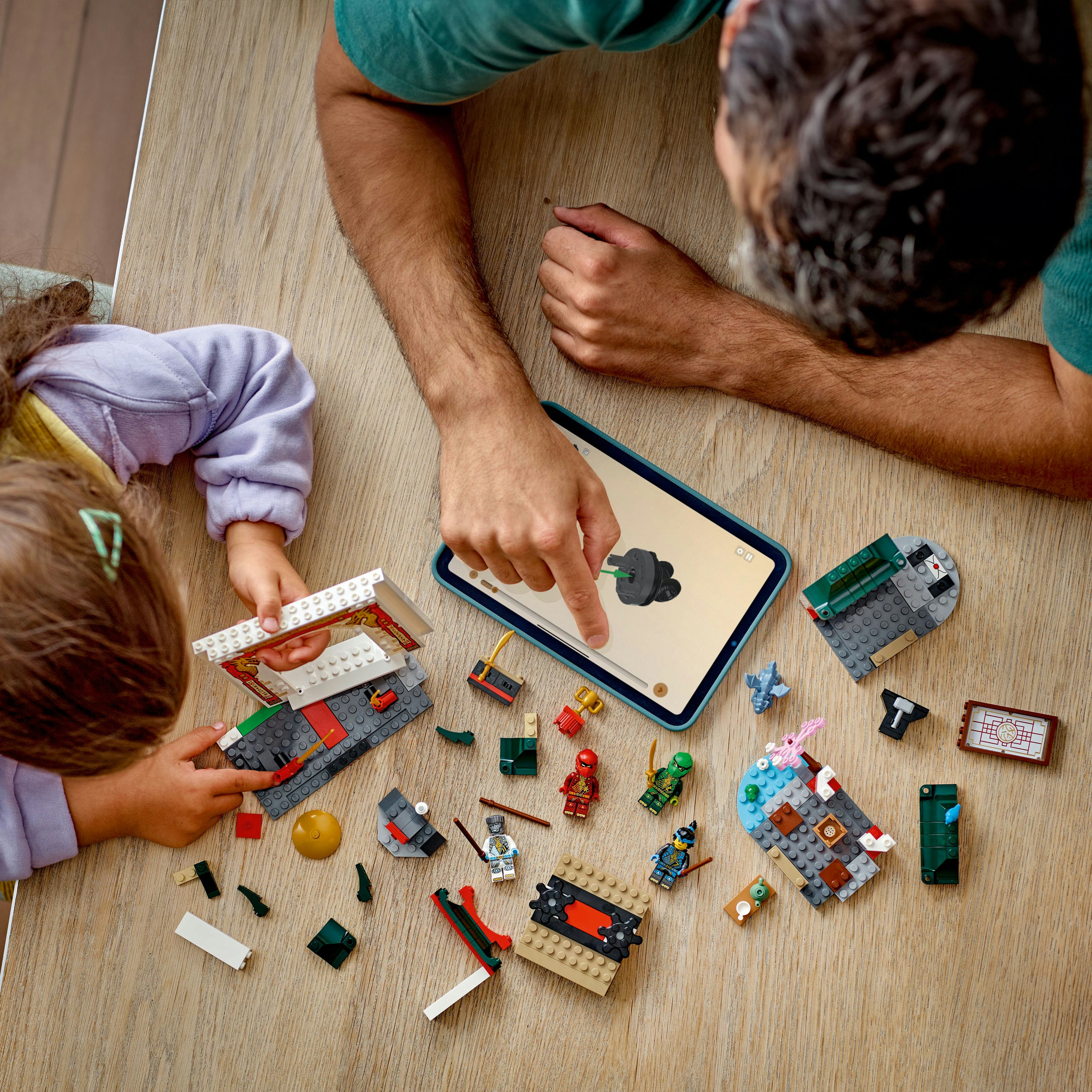 Dziecko i dorosły budują Lego na drewnie. Tablet pokazuje instrukcje. Klocki Lego i figurki są rozrzucone.