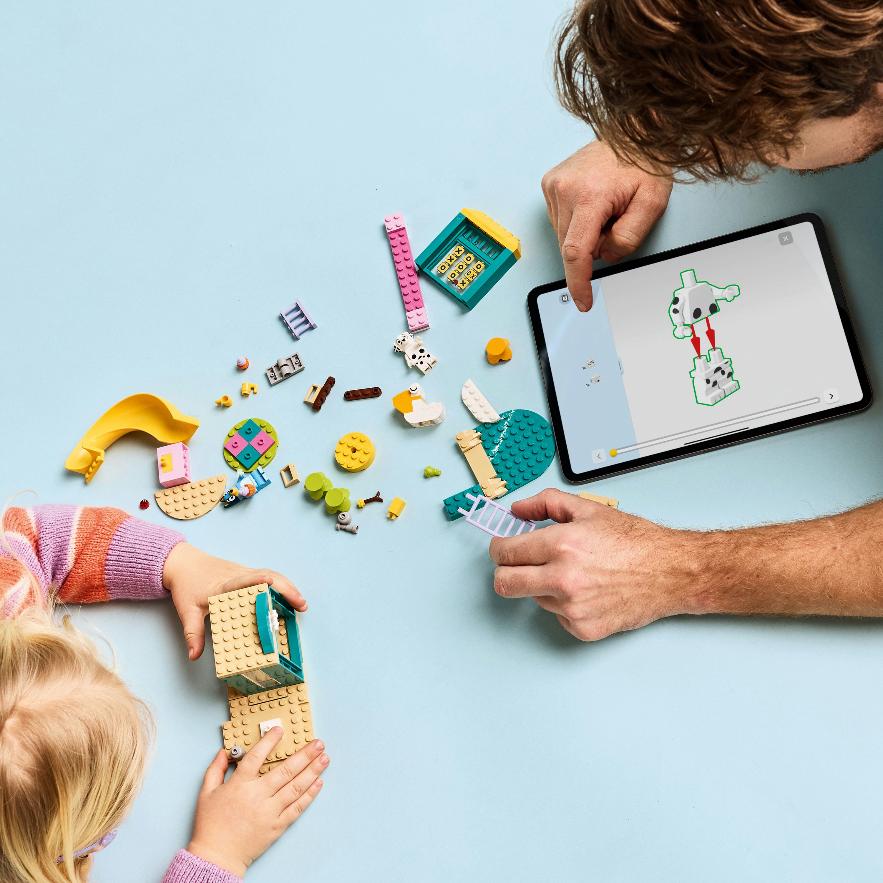 Dziecko i dorosły budują z klocków LEGO, korzystając z instrukcji na tablecie. Różne elementy są rozrzucone na niebieskiej powierzchni.