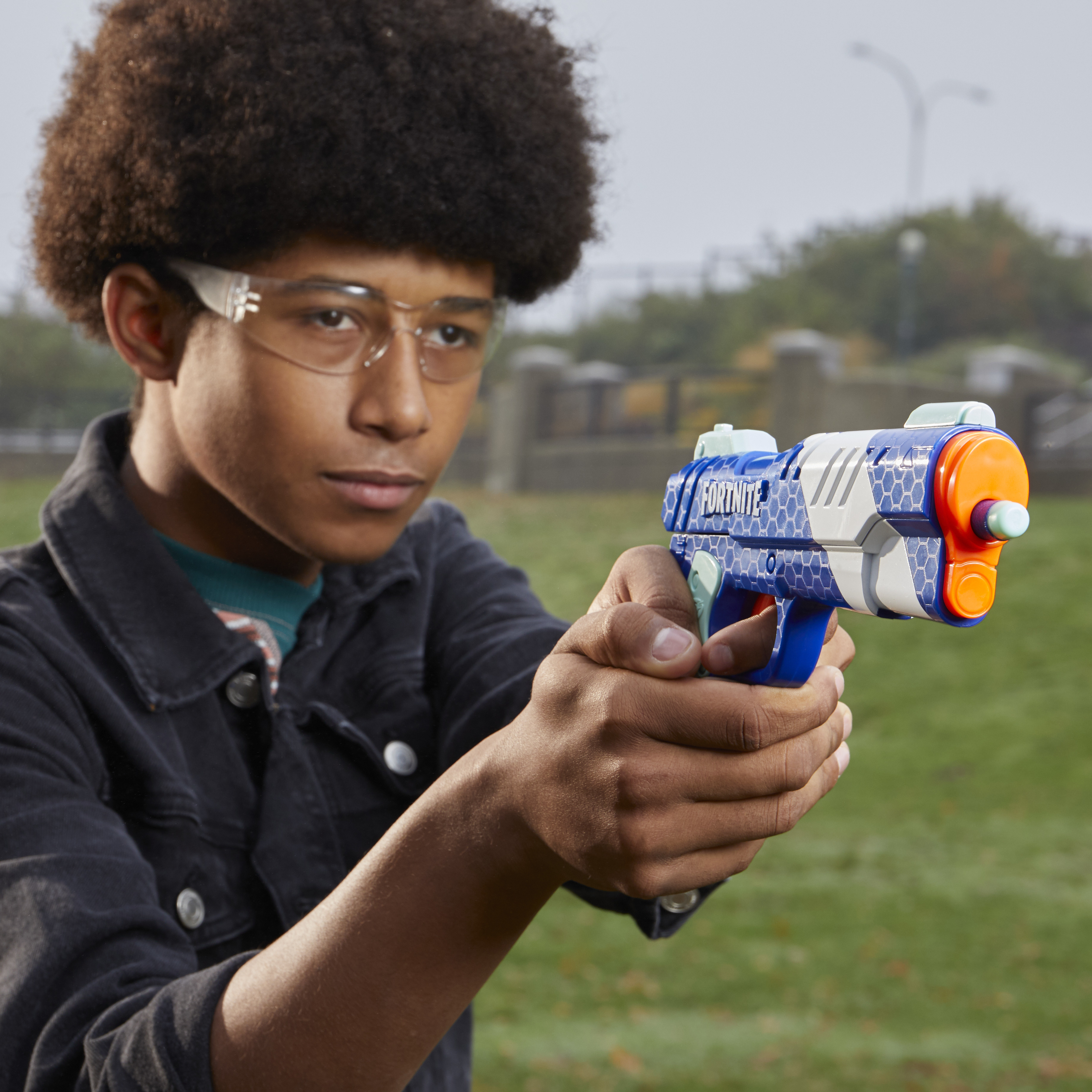 Chłopiec w okularach trzyma niebiesko-pomarańczowy pistolet-zabawkę. Stoi przed zieloną murawą i budynkiem.
