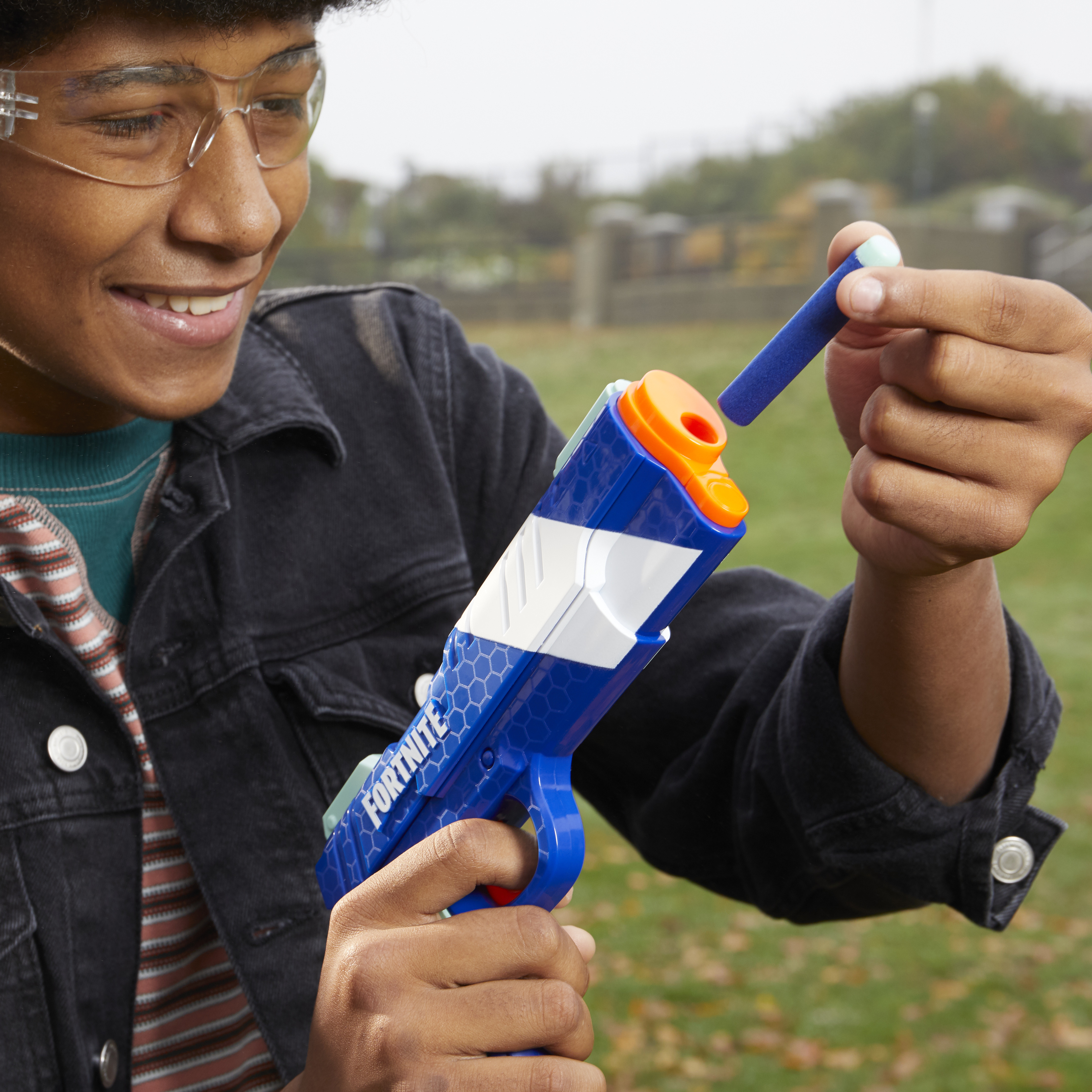Osoba trzyma niebiesko-pomarańczową zabawkę-pistolet, wkładając piankową strzałkę. Osoba nosi okulary i czarną kurtkę.