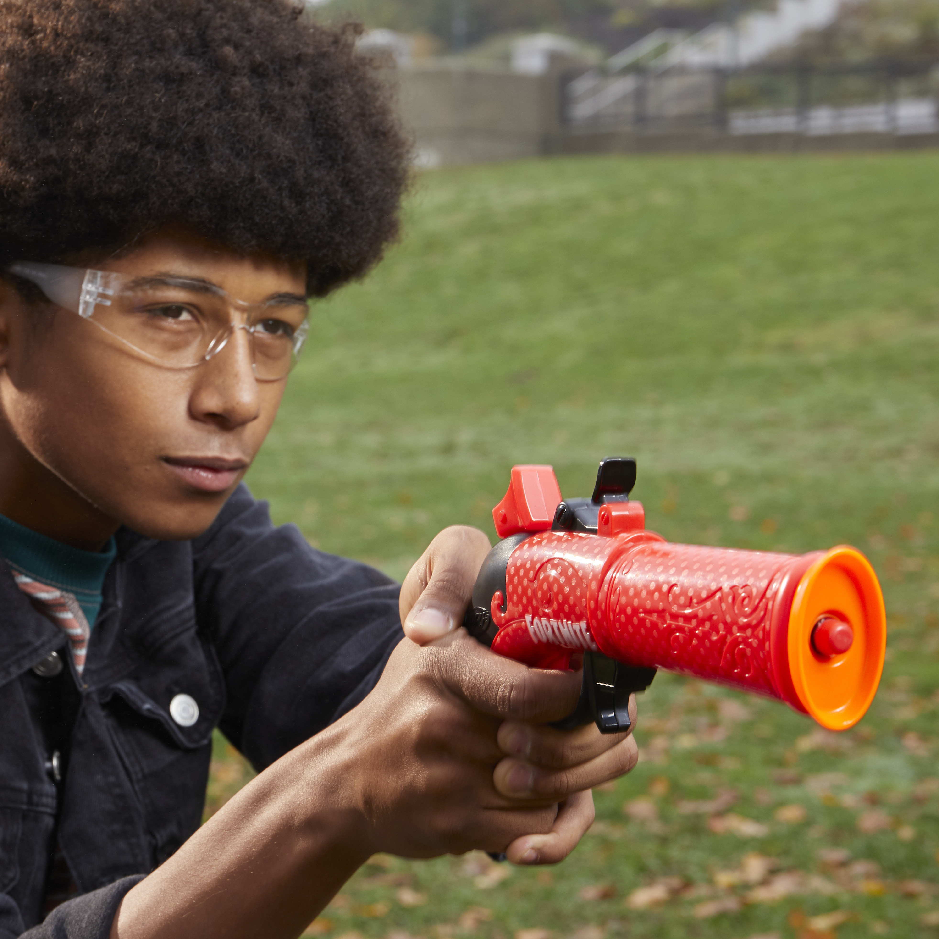 Nastolatek trzyma czerwoną i pomarańczową zabawkę-pistolet, w okularach ochronnych. Tło: zielony trawnik i liście.