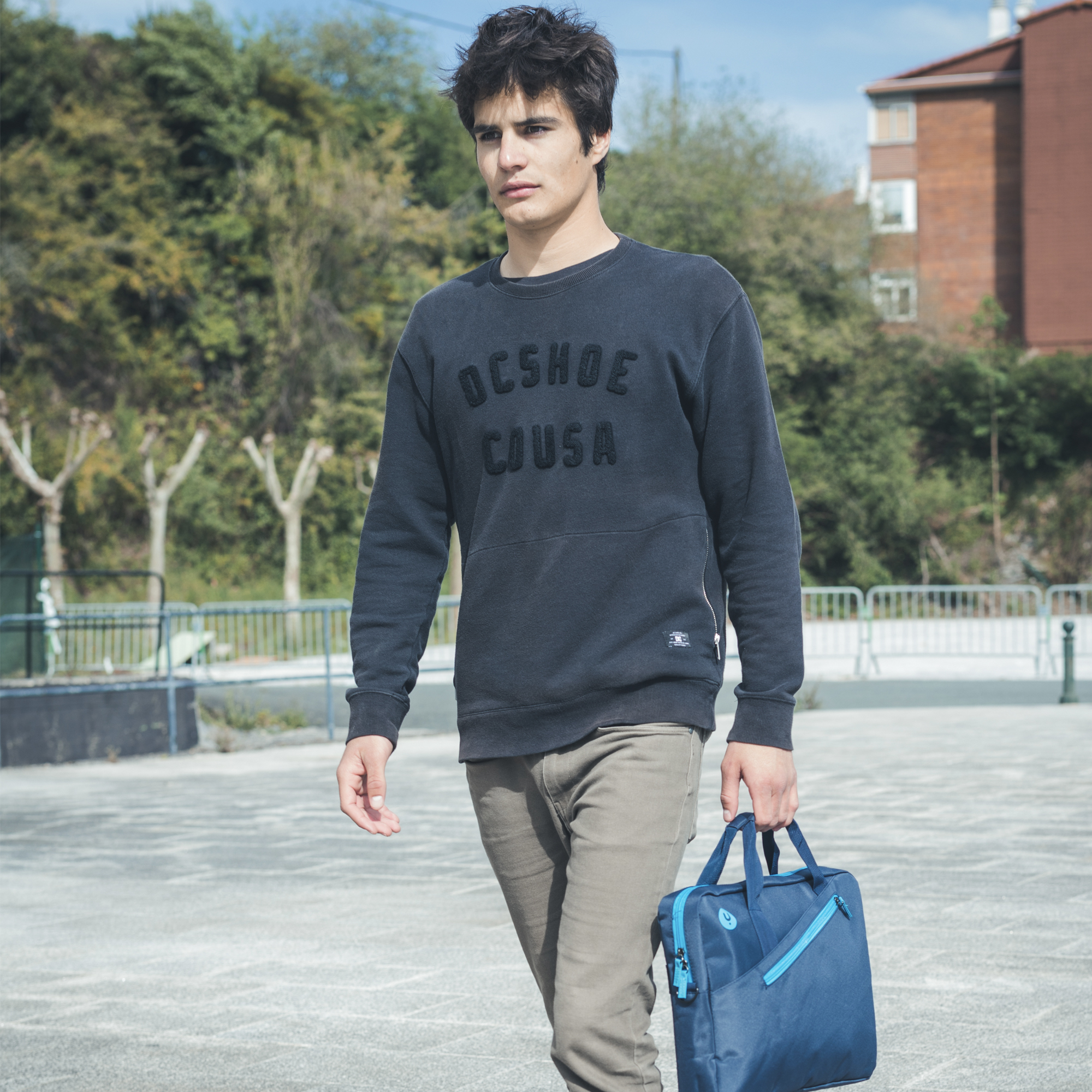 Ein junger Mann geht mit einer blauen Tasche. Er trägt einen dunklen Pullover, olivfarbene Hose. Bäume und ein Gebäude im Hintergrund.