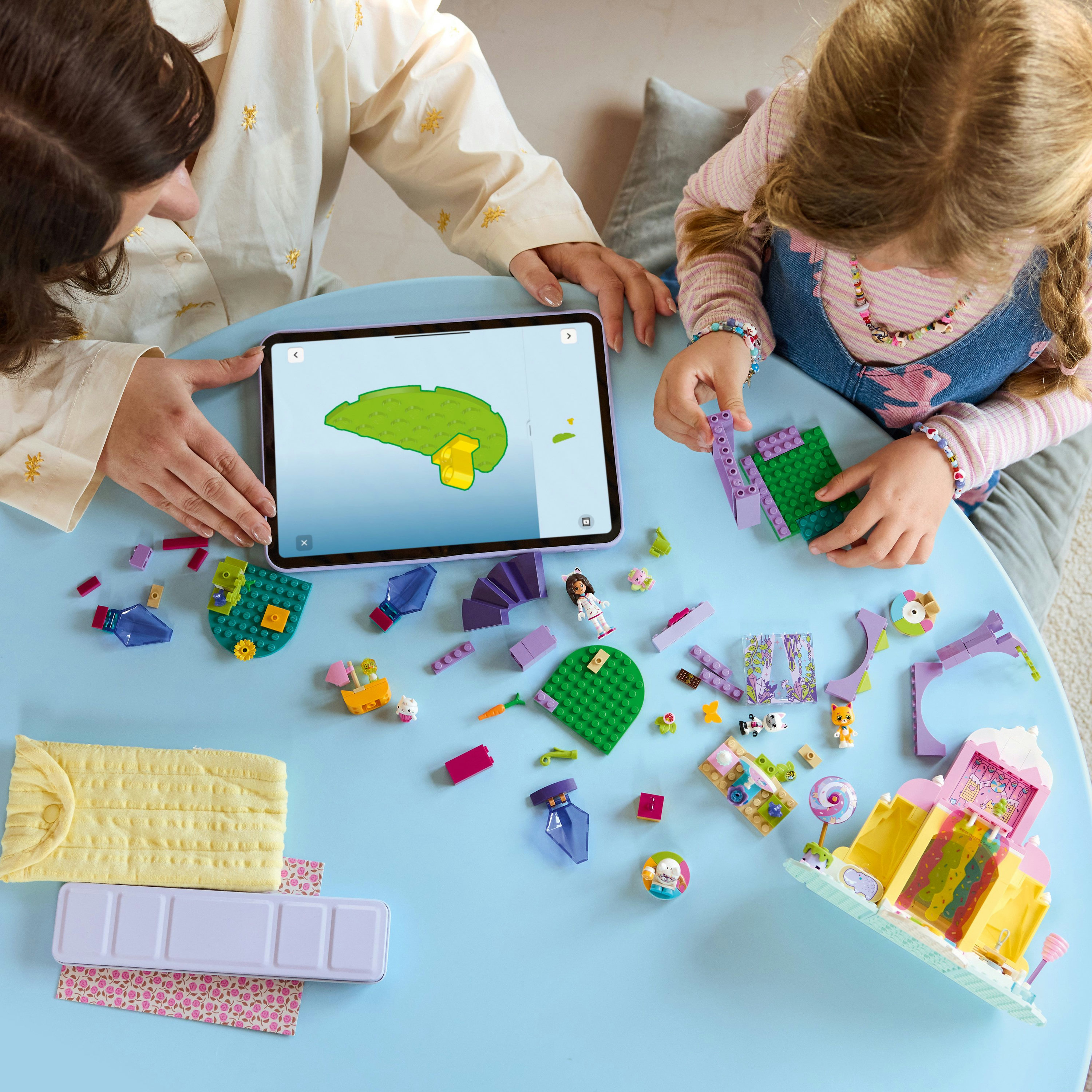 Dwie osoby budują z kolorowych klocków na niebieskim stole. Jedna patrzy na tablet.