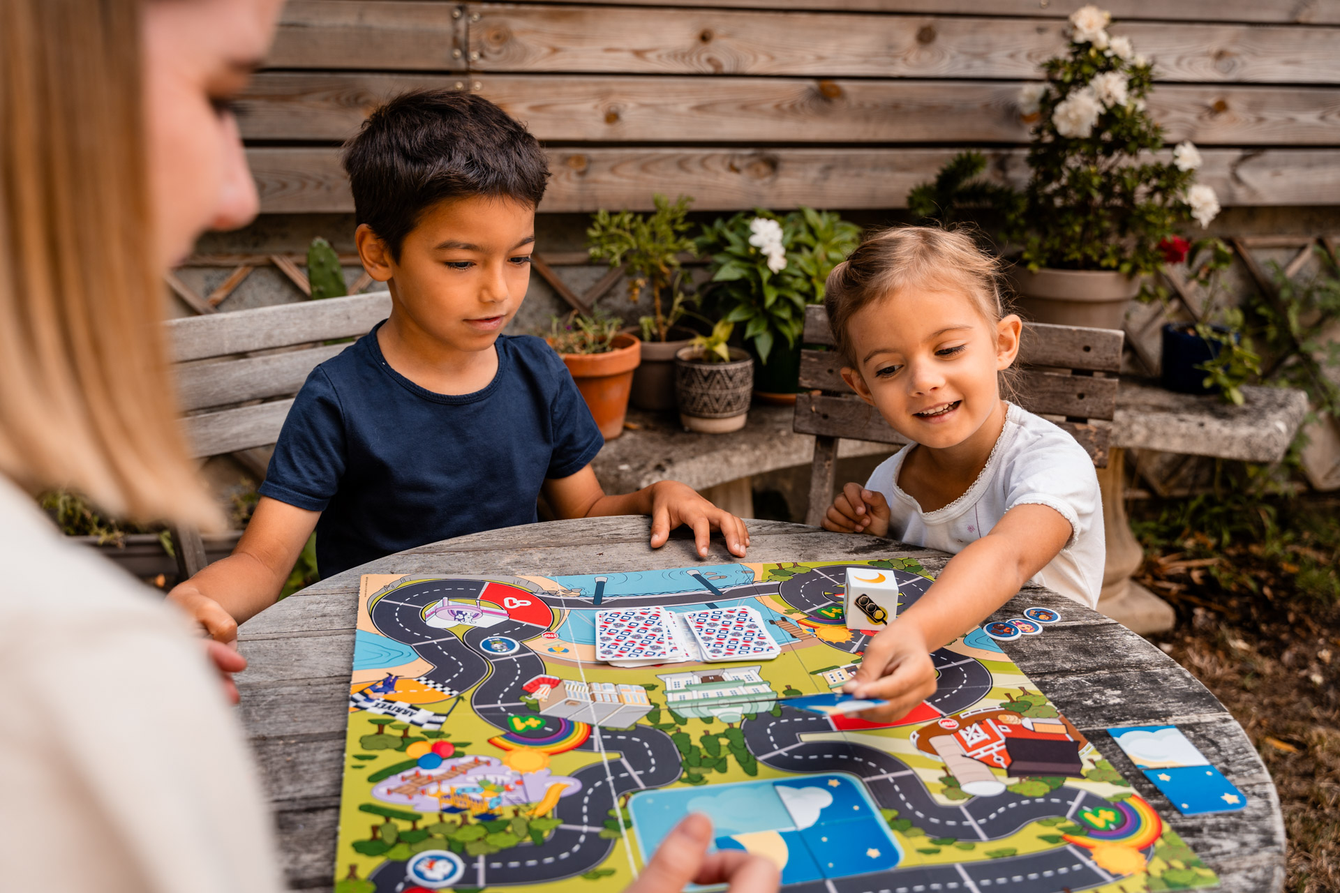 Tre bambini giocano a un gioco da tavolo all'aperto su un tavolo di legno. Sullo sfondo una staccionata di legno e piante.