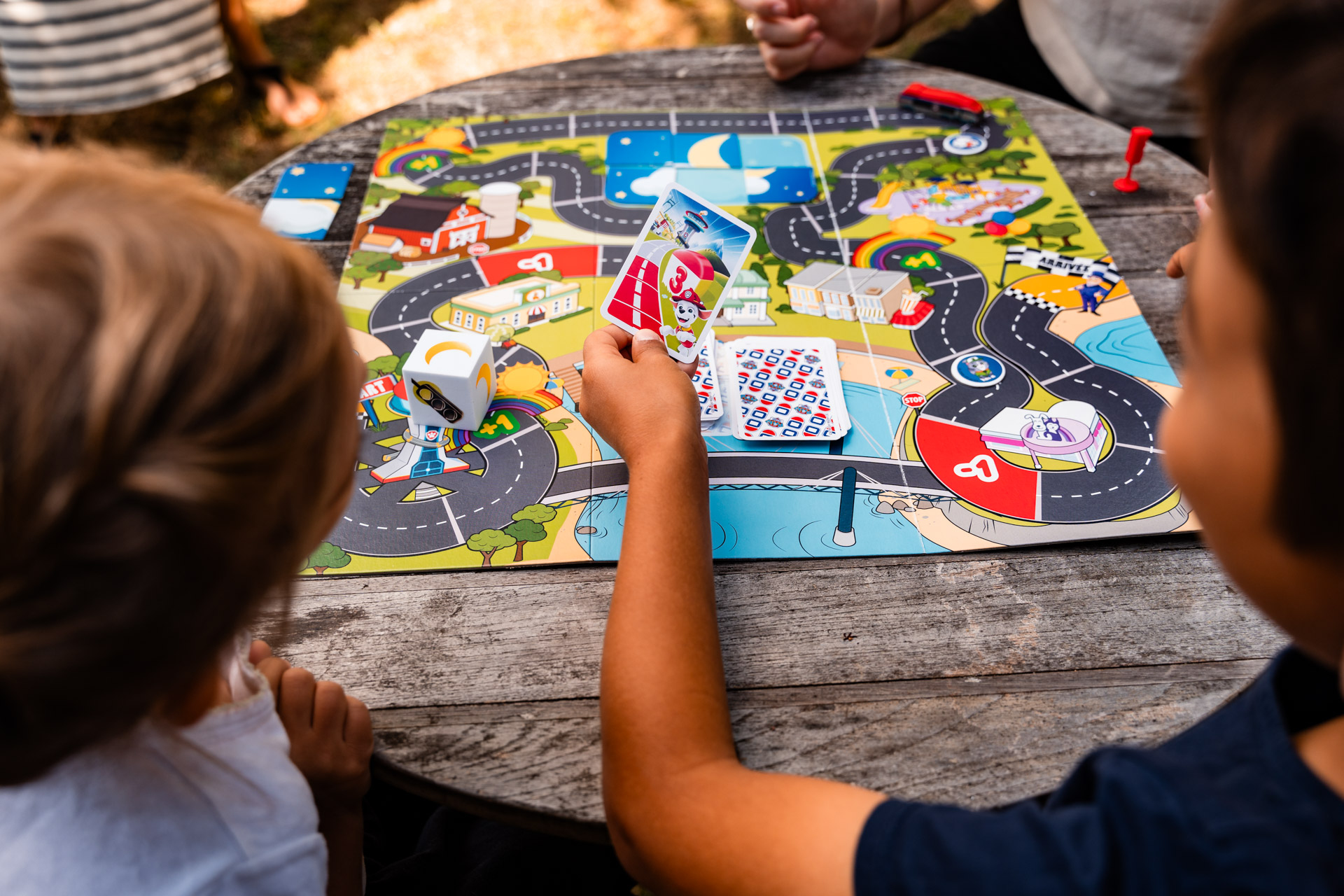 Bambini giocano a un gioco da tavolo su un tavolo di legno. Un bambino tiene carte Paw Patrol. Il tabellone è colorato.