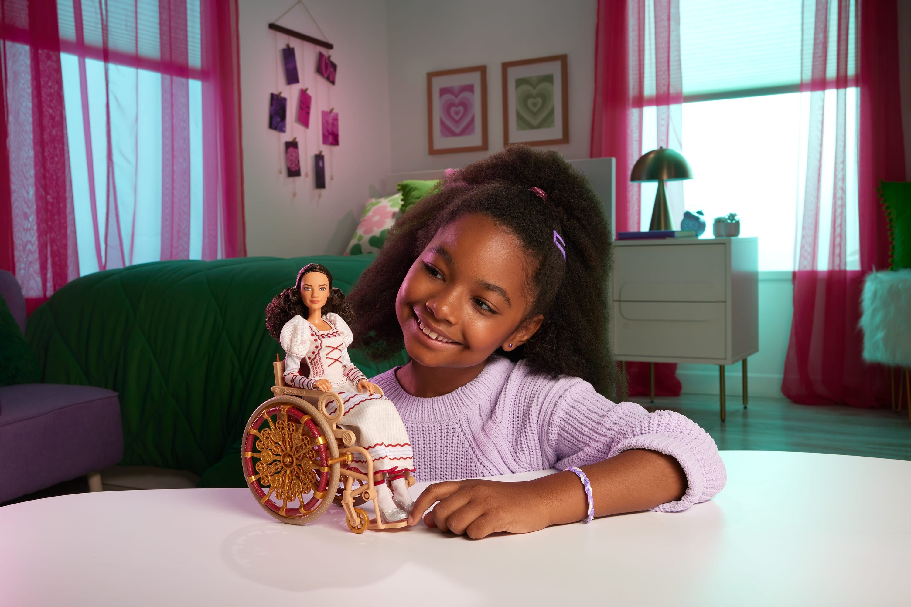 Dziewczyna uśmiecha się do lalki na wózku inwalidzkim. Różowe zasłony i łóżko w tle.