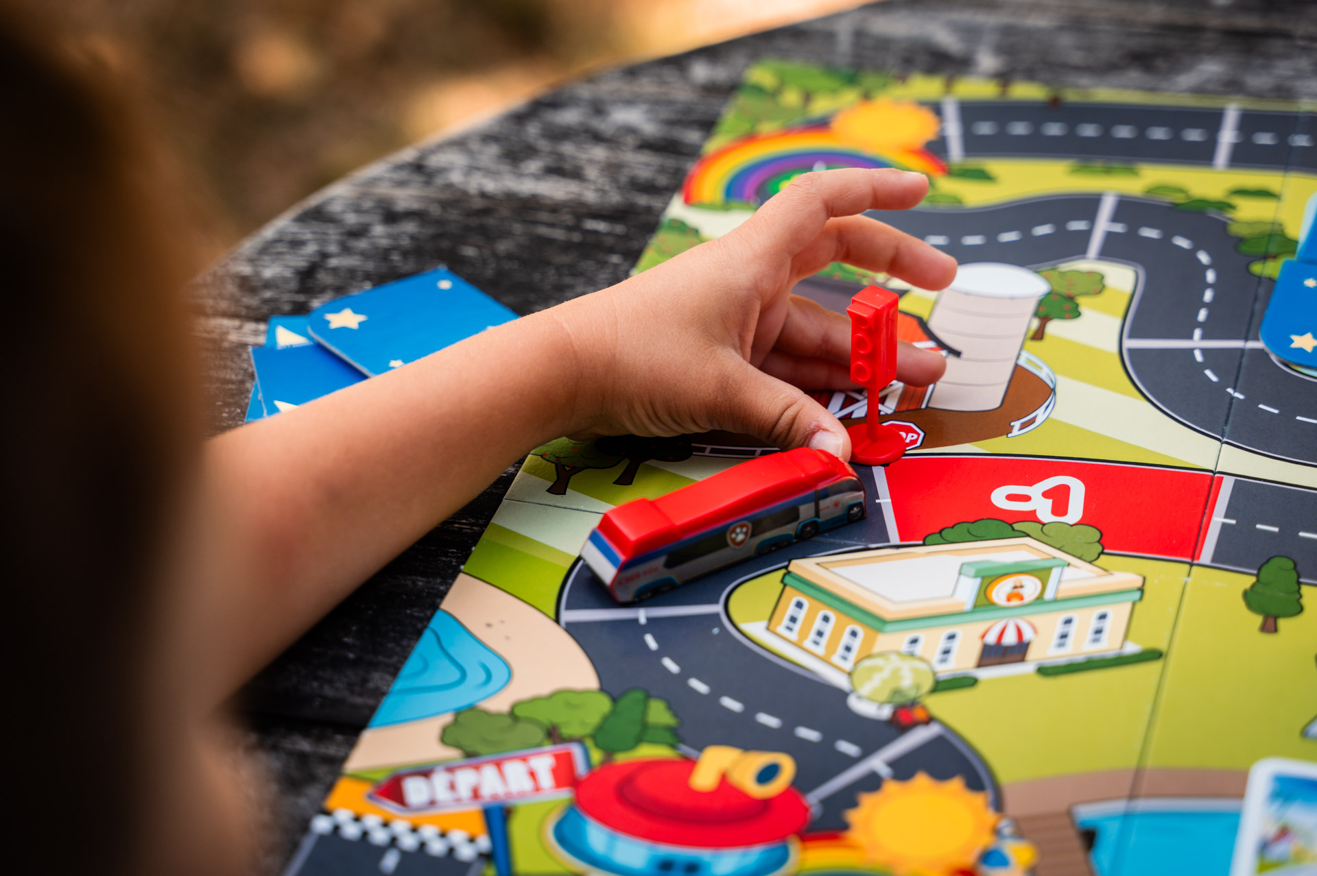 La mano di un bambino posiziona un autobus giocattolo su un tabellone. Il tabellone mostra strade, edifici e un arcobaleno. Un semaforo rosso.