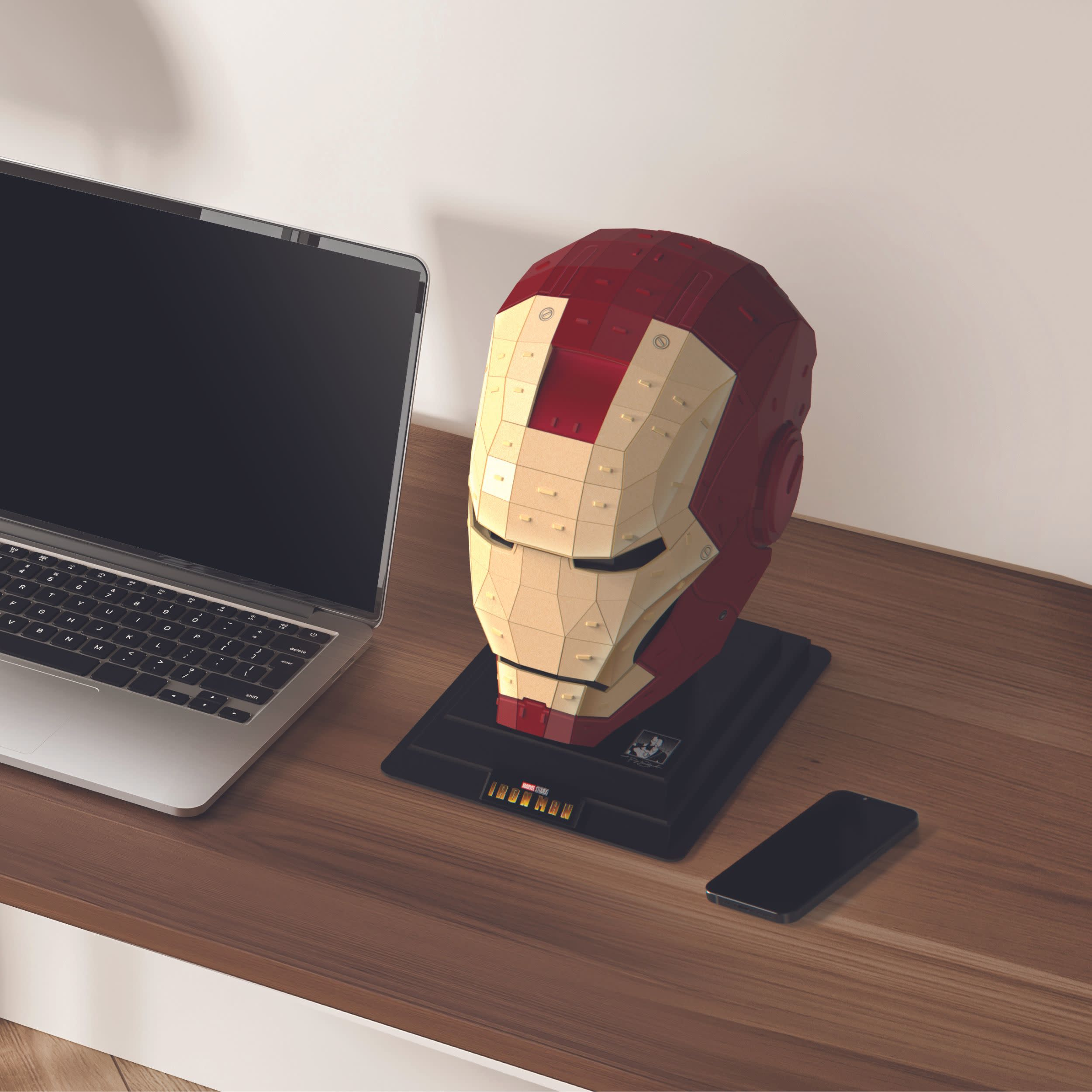 Iron-Man-Helm auf einem Ständer, Laptop und Telefon auf einem Holzschreibtisch. Weiße Wand im Hintergrund.