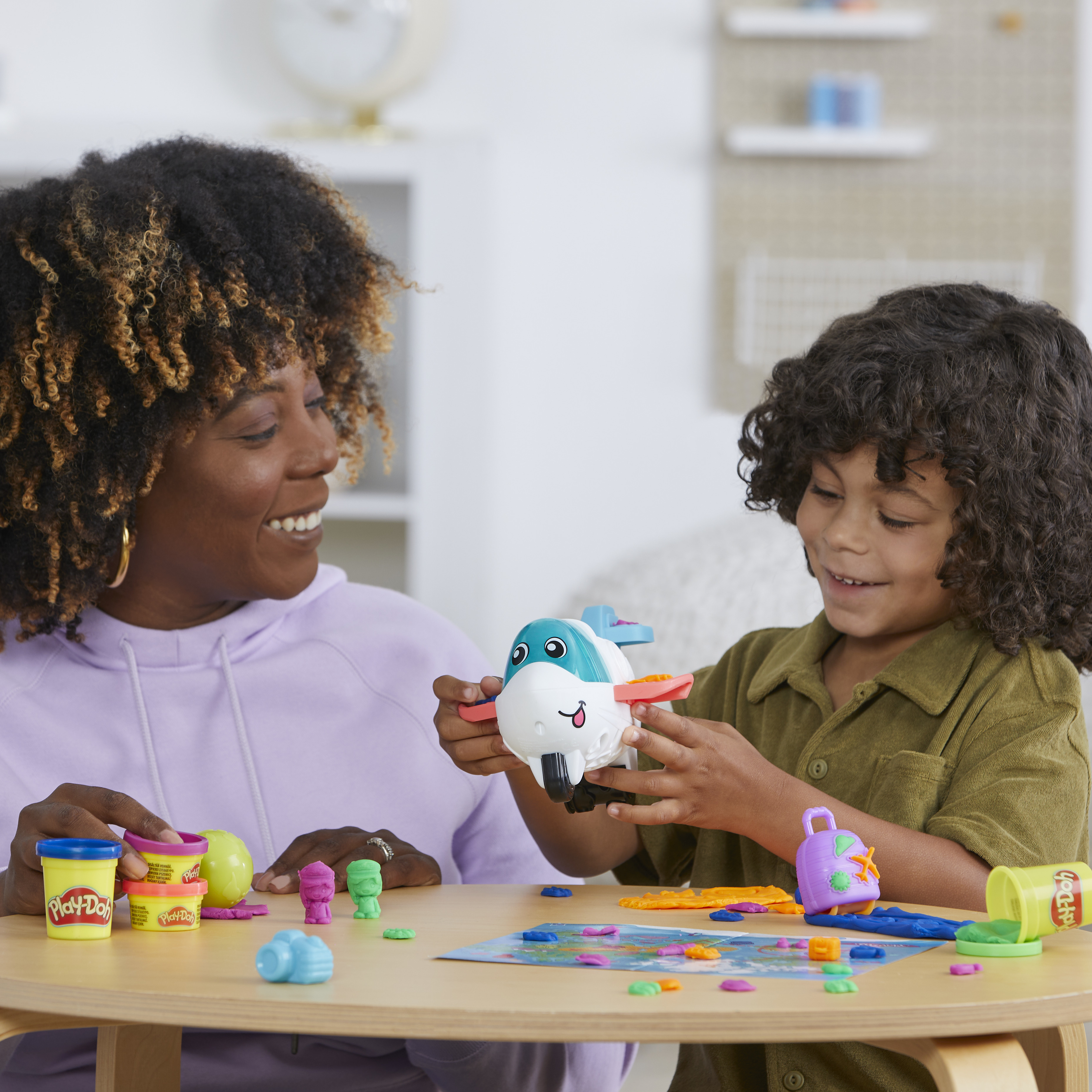 Una mujer y un niño juegan con Play-Doh y un avión de juguete. Hay botes y formas de Play-Doh de colores sobre la mesa.
