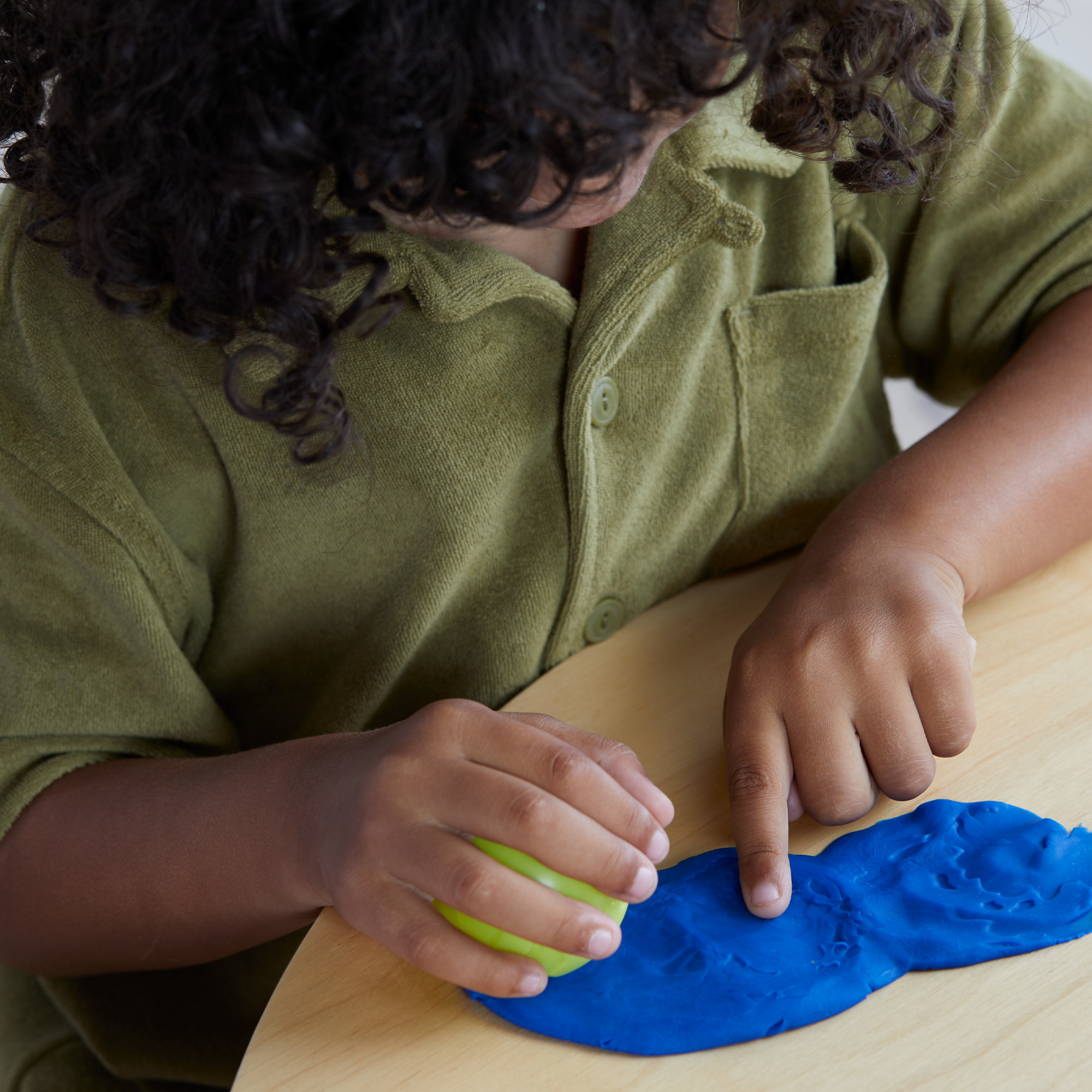 Un niño con camisa verde juega con plastilina azul y verde sobre una mesa de madera clara. Se ve pelo rizado.