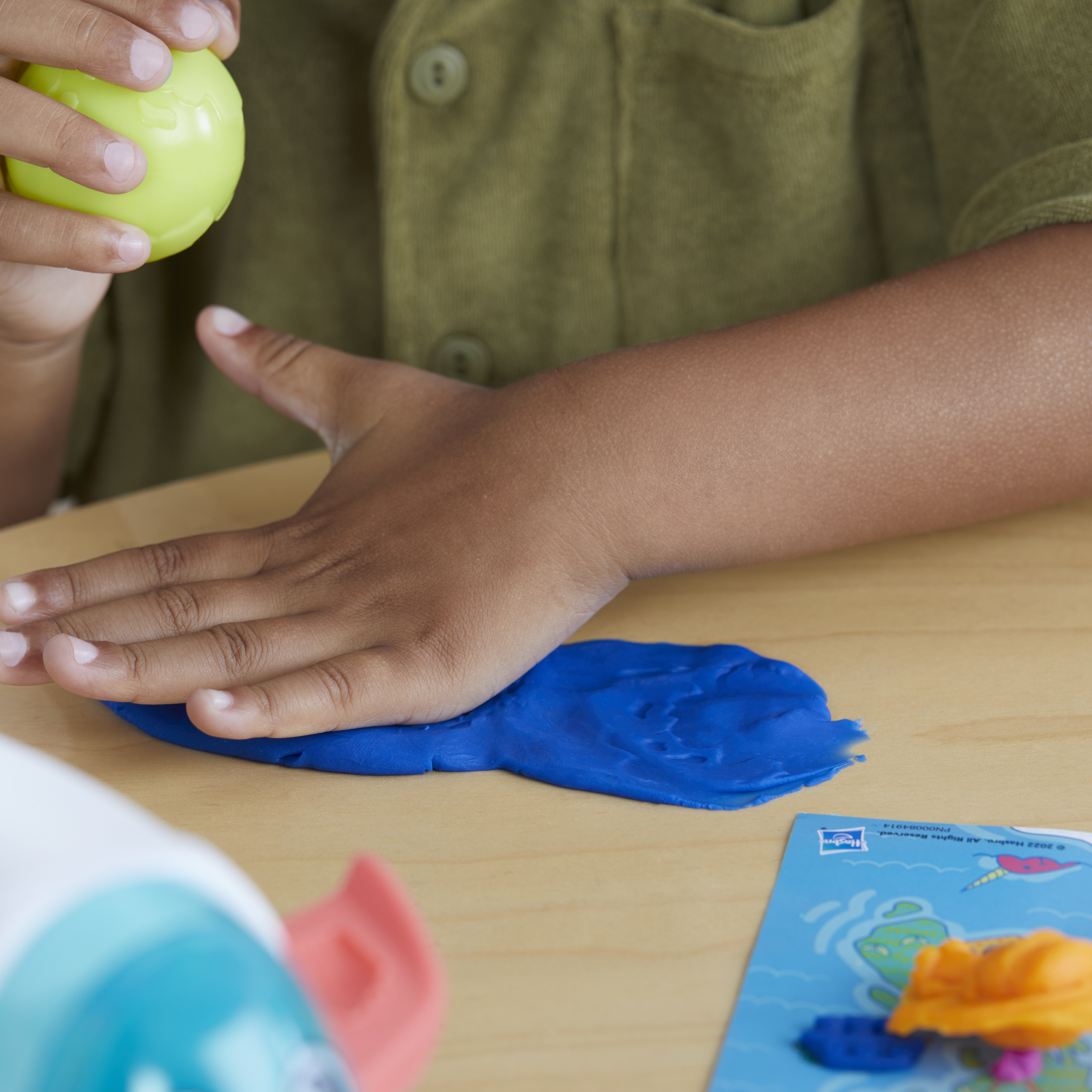 Un niño presiona plastilina azul con una mano. Una pelota amarilla se sostiene en la otra mano. Un juguete y una camisa son visibles.