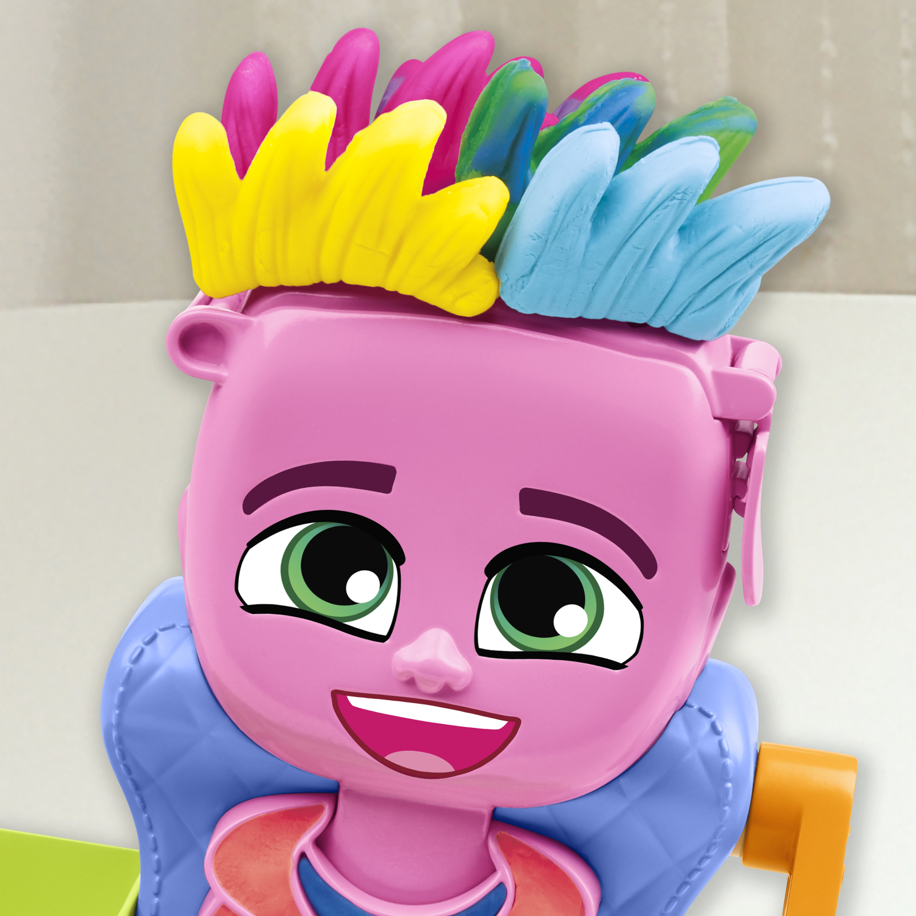 Juguete rosa con pelo en forma de pluma, ojos verdes y boca sonriente. Se ven detalles azules y naranjas.