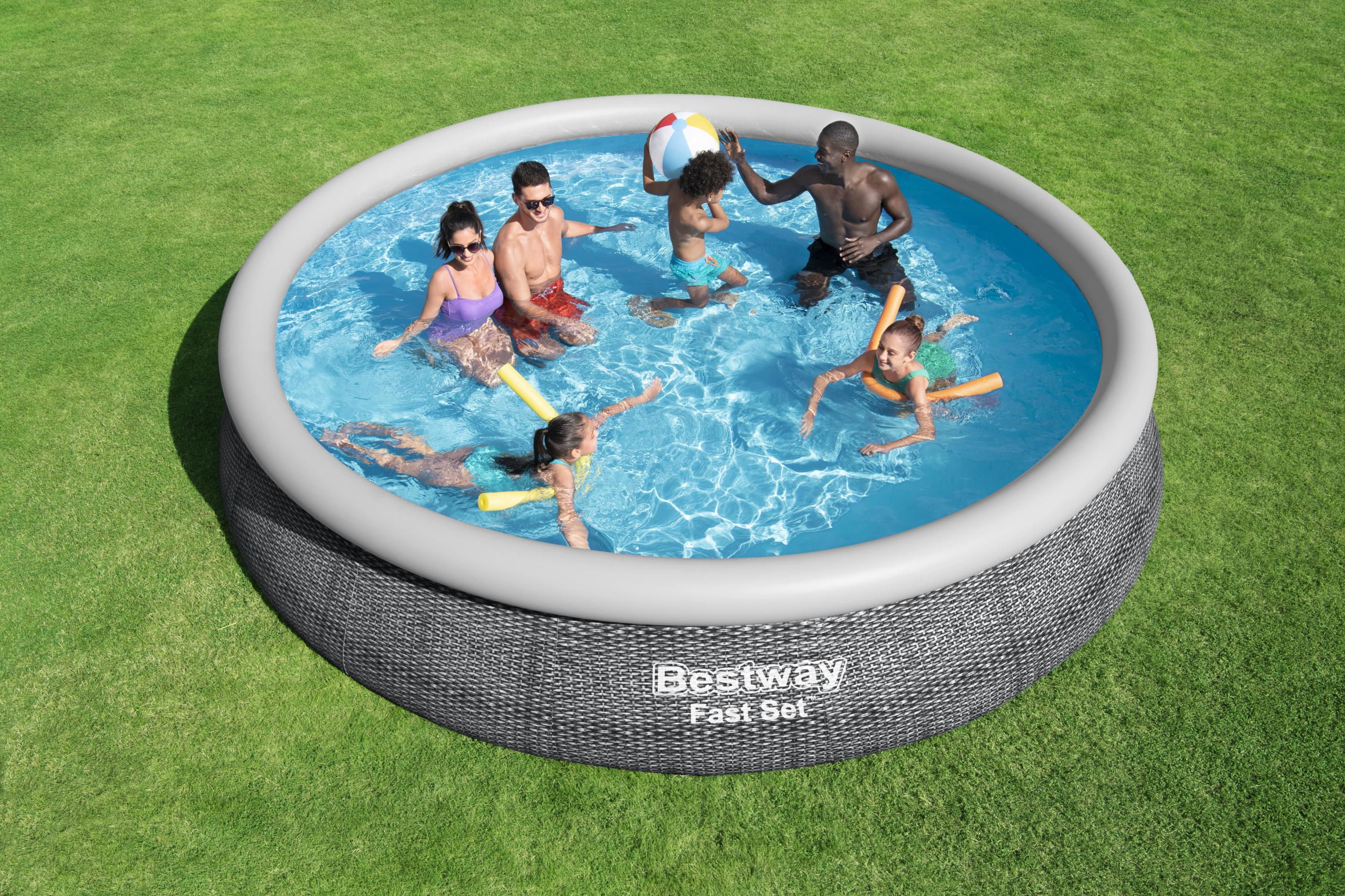 Menschen in einem Pool, einige mit Schwimmnudeln und einem Ball, auf einem grünen Rasen. Der Pool ist rund und grau.