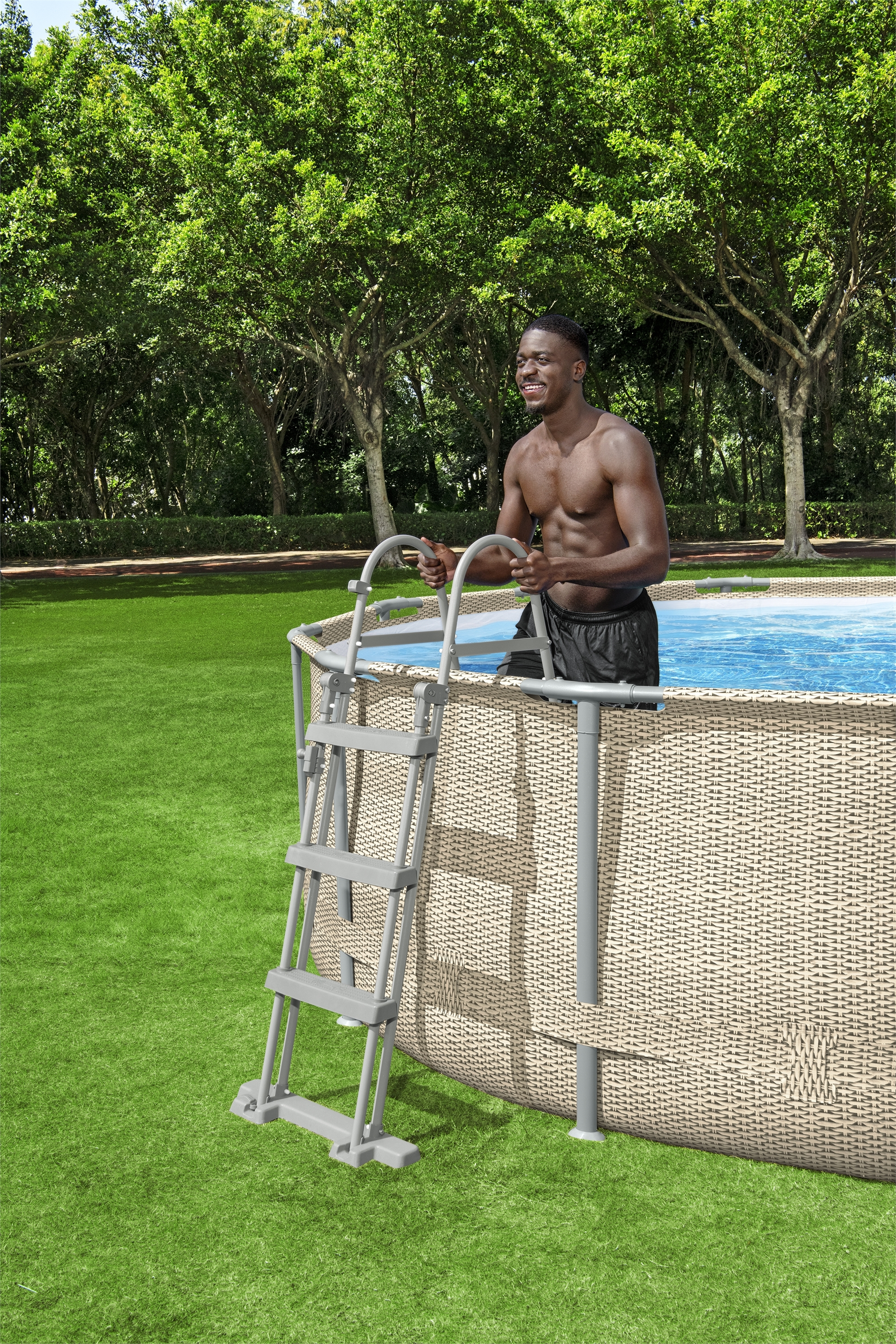 Un hombre en pantalones cortos negros sonríe, sosteniendo una escalera de piscina. La piscina está detrás de él. Hay árboles y hierba verde en el fondo.