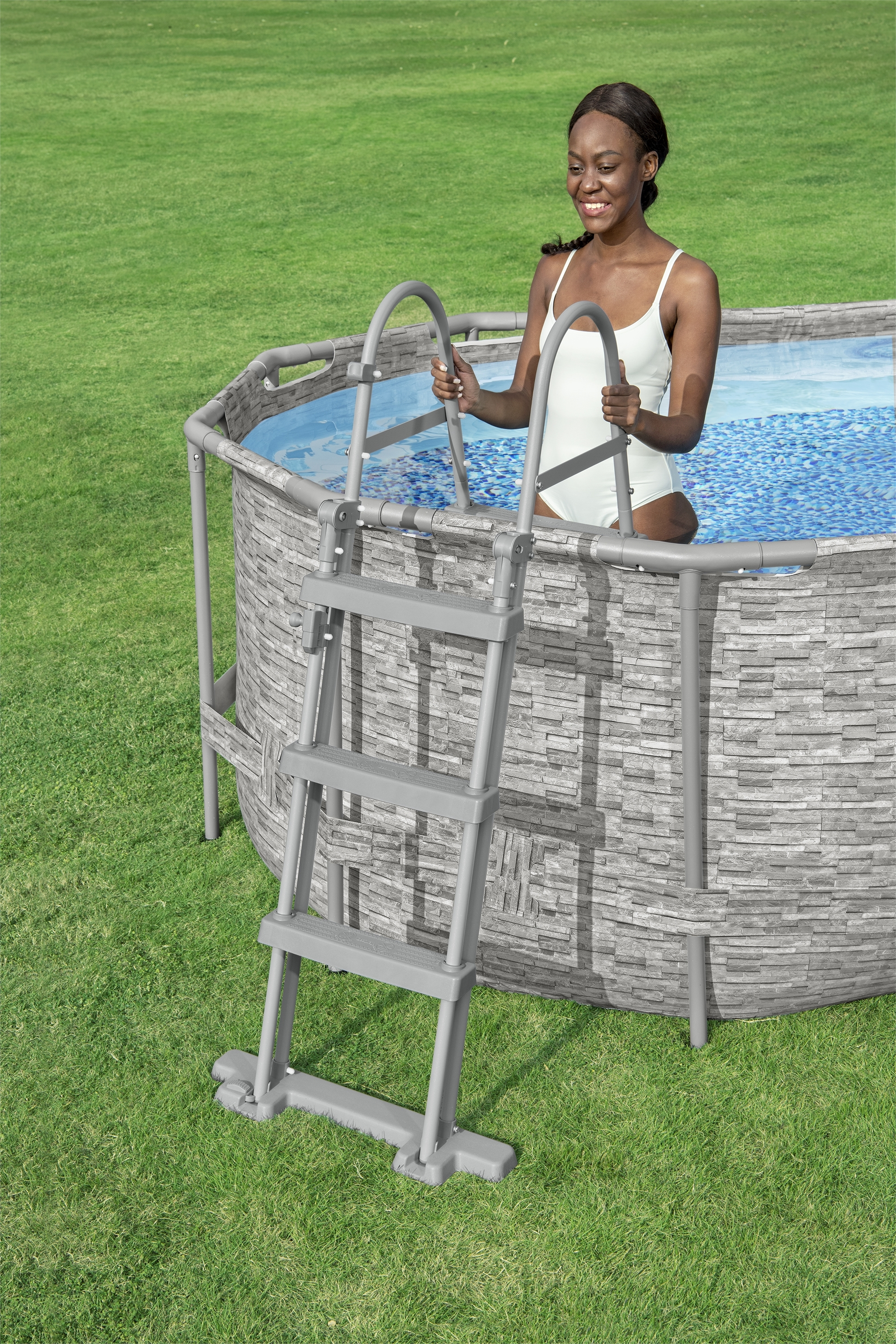 Una mujer con traje de baño blanco está en una piscina, agarrando la escalera. La piscina está en un jardín.