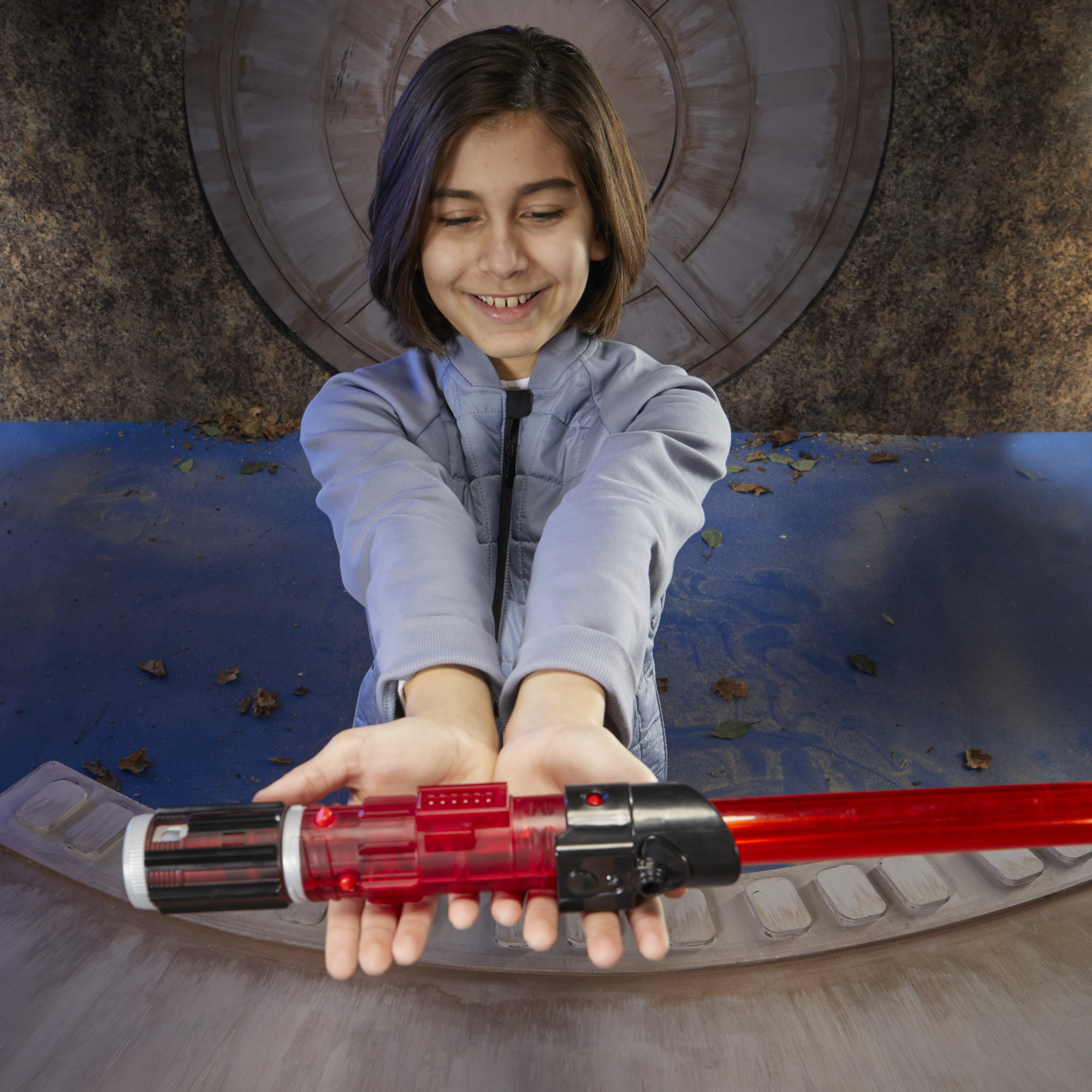 Uśmiechnięta osoba trzyma czerwony miecz świetlny. Mają na sobie szarą kurtkę. Tło ma brązową ścianę i niebieską powierzchnię.