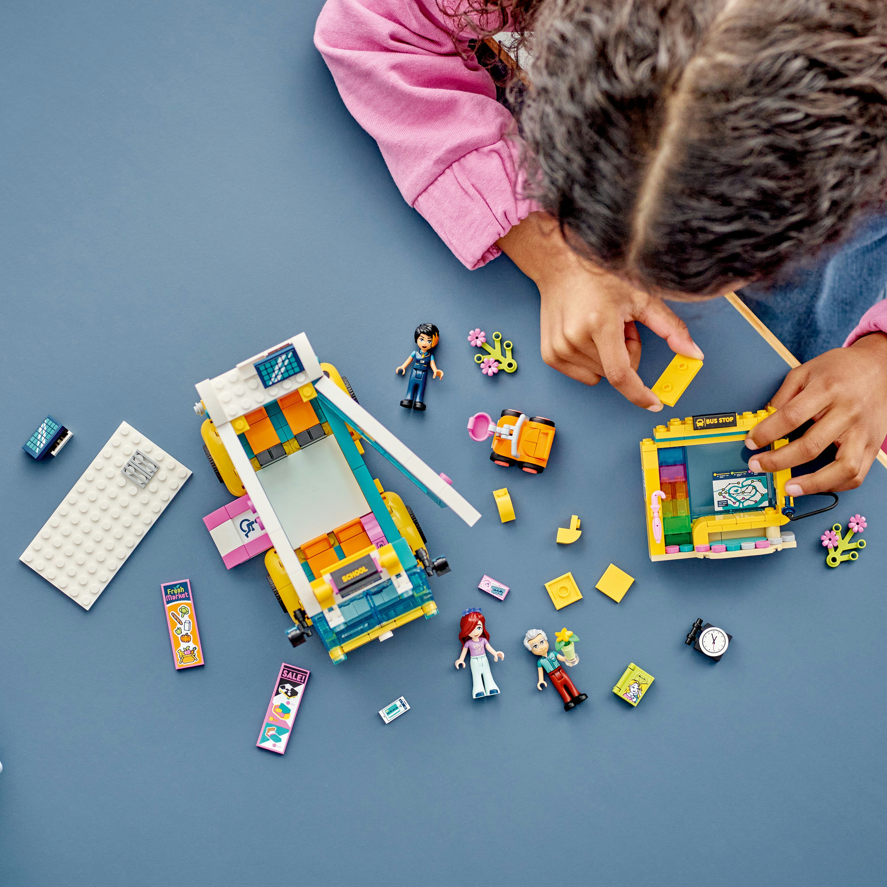 Dziecko bawi się klockami Lego, składając autobus i przystanek autobusowy, figurki i różne kolorowe elementy.