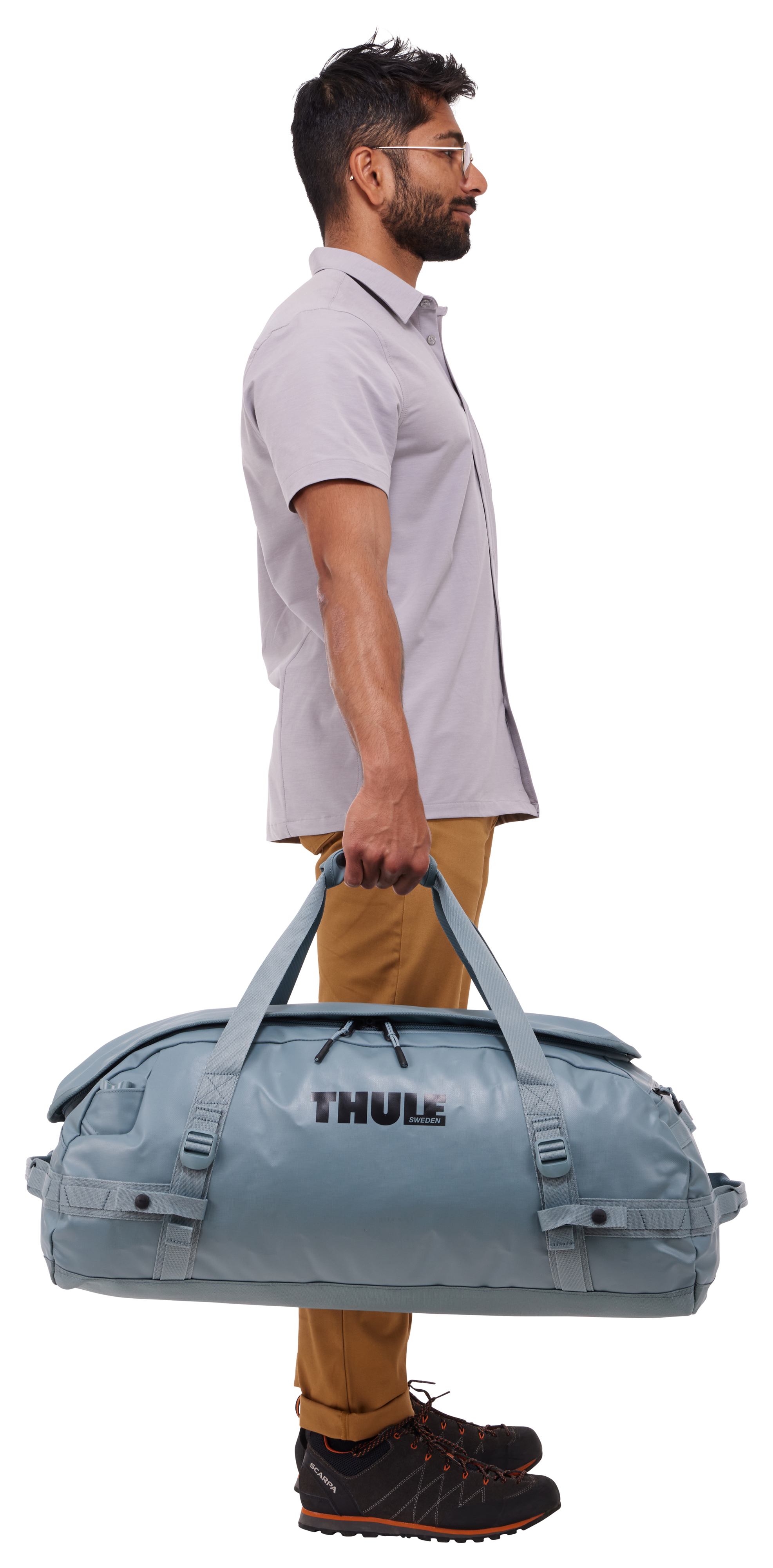 Ein Mann in hellem Hemd und Hose hält eine blaue Thule-Tasche. Er trägt eine Brille und schwarze Schuhe. Weißer Hintergrund.