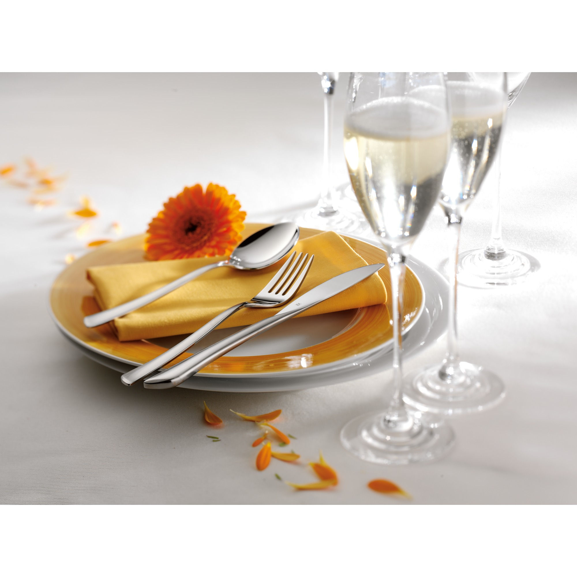 Ein gedeckter Tisch mit Besteck, einer gelben Serviette, einer orangefarbenen Blume, Tellern und Sektgläsern, mit Blütenblatt-Akzenten.