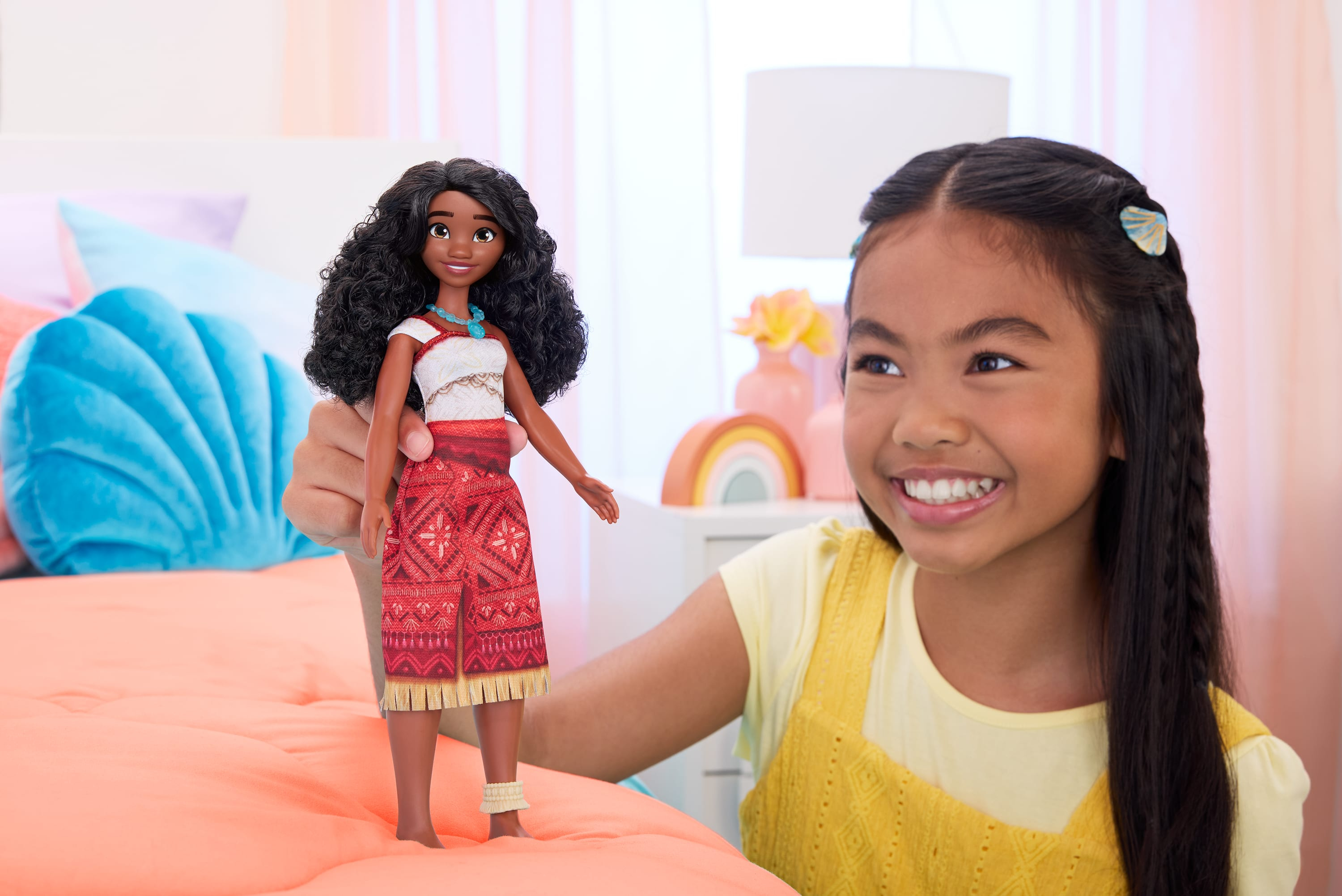 Ein Mädchen hält eine Moana-Puppe. Das Mädchen lächelt. Im Hintergrund befindet sich ein Bett mit Kissen.