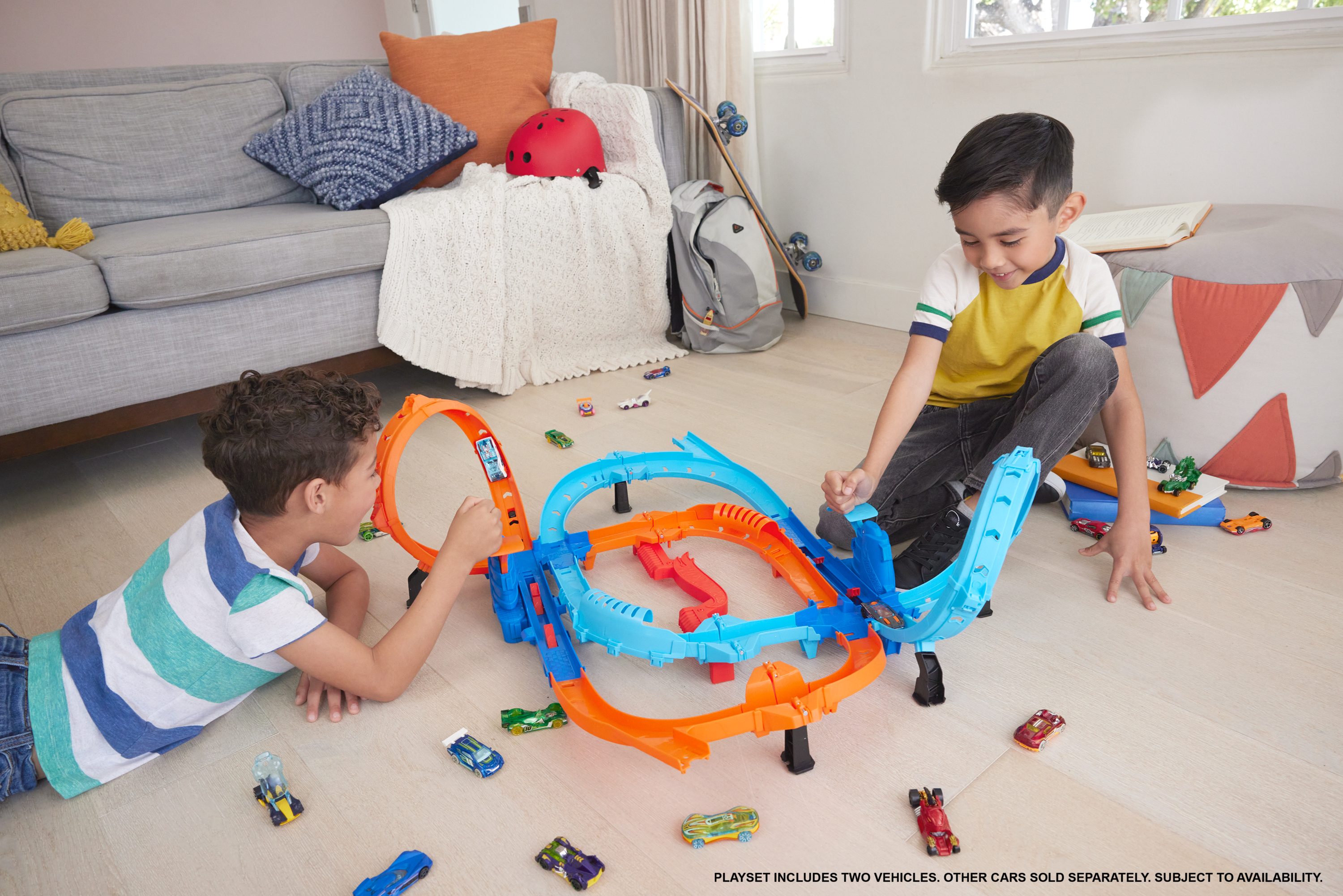 Zwei Jungen spielen mit einer Spielzeugauto-Rennstrecke. Autos sind verstreut. Im Hintergrund sind ein Sofa und ein Skateboard.