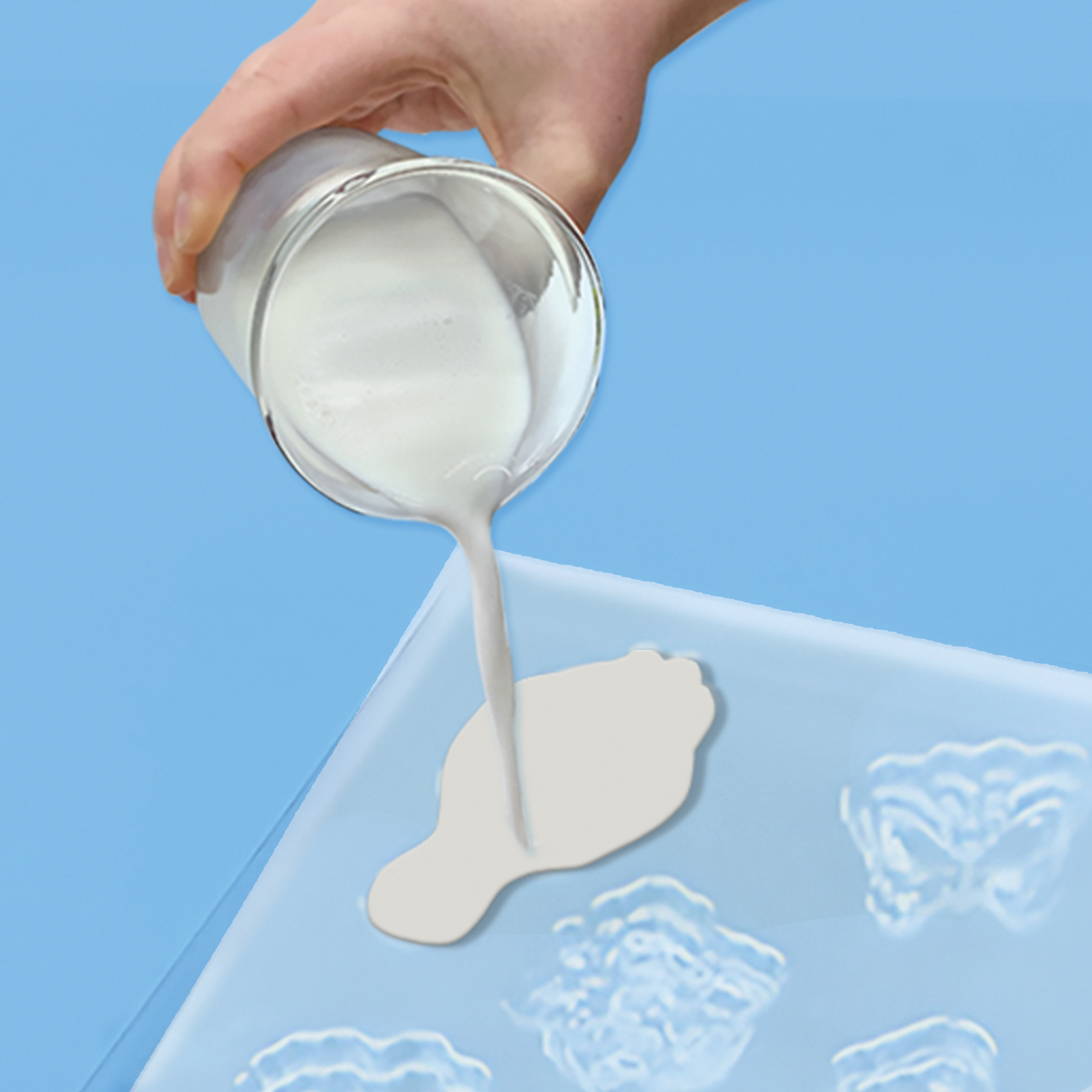 Ręka wlewa białą ciecz ze szklanki do niebieskiej formy. Forma ma kilka kształtów. Niebieskie tło.