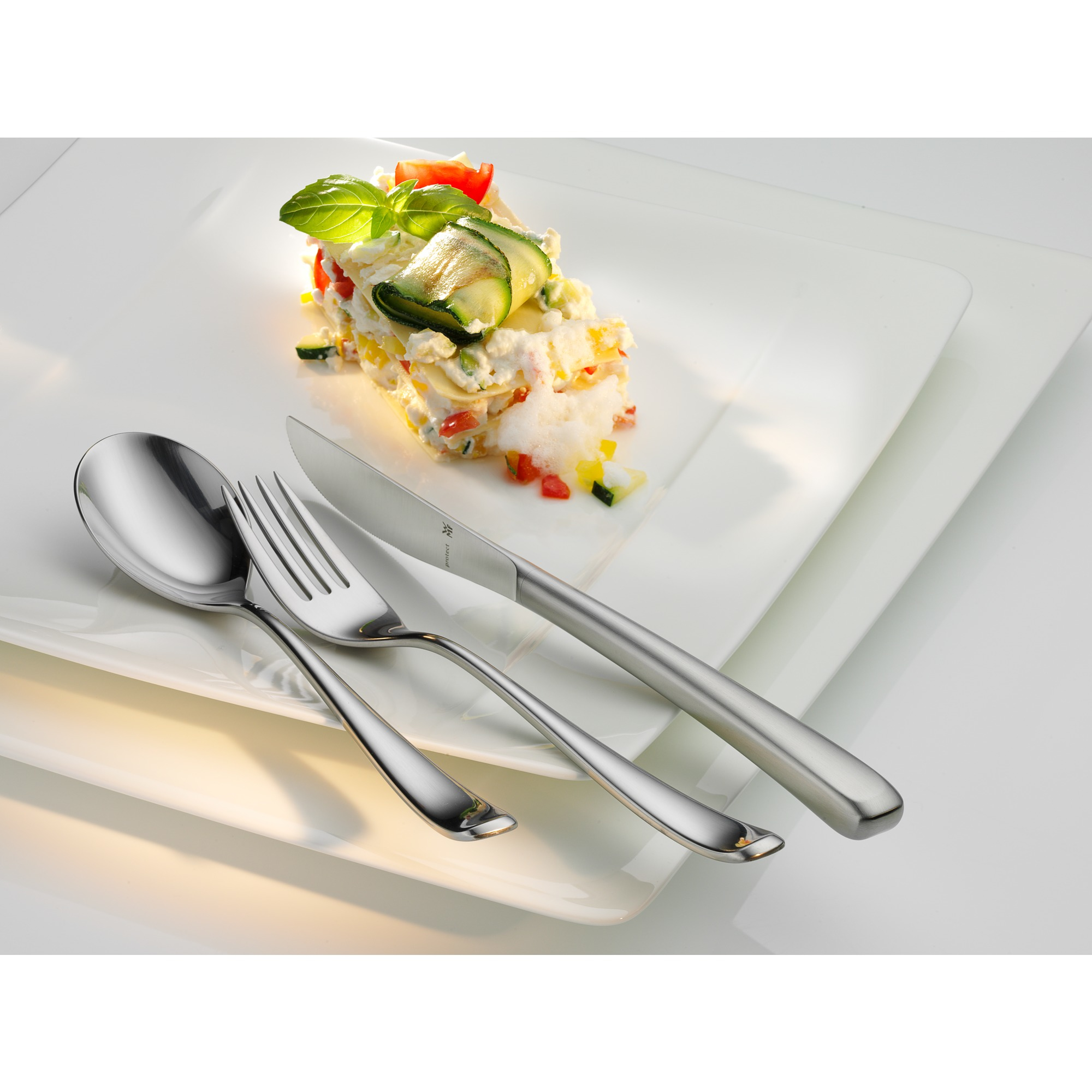 Ein Teller mit Essen, Besteck und zwei weiteren Tellern darunter. Das Essen ist ein geschichtetes Gericht mit Gemüse und Basilikum.