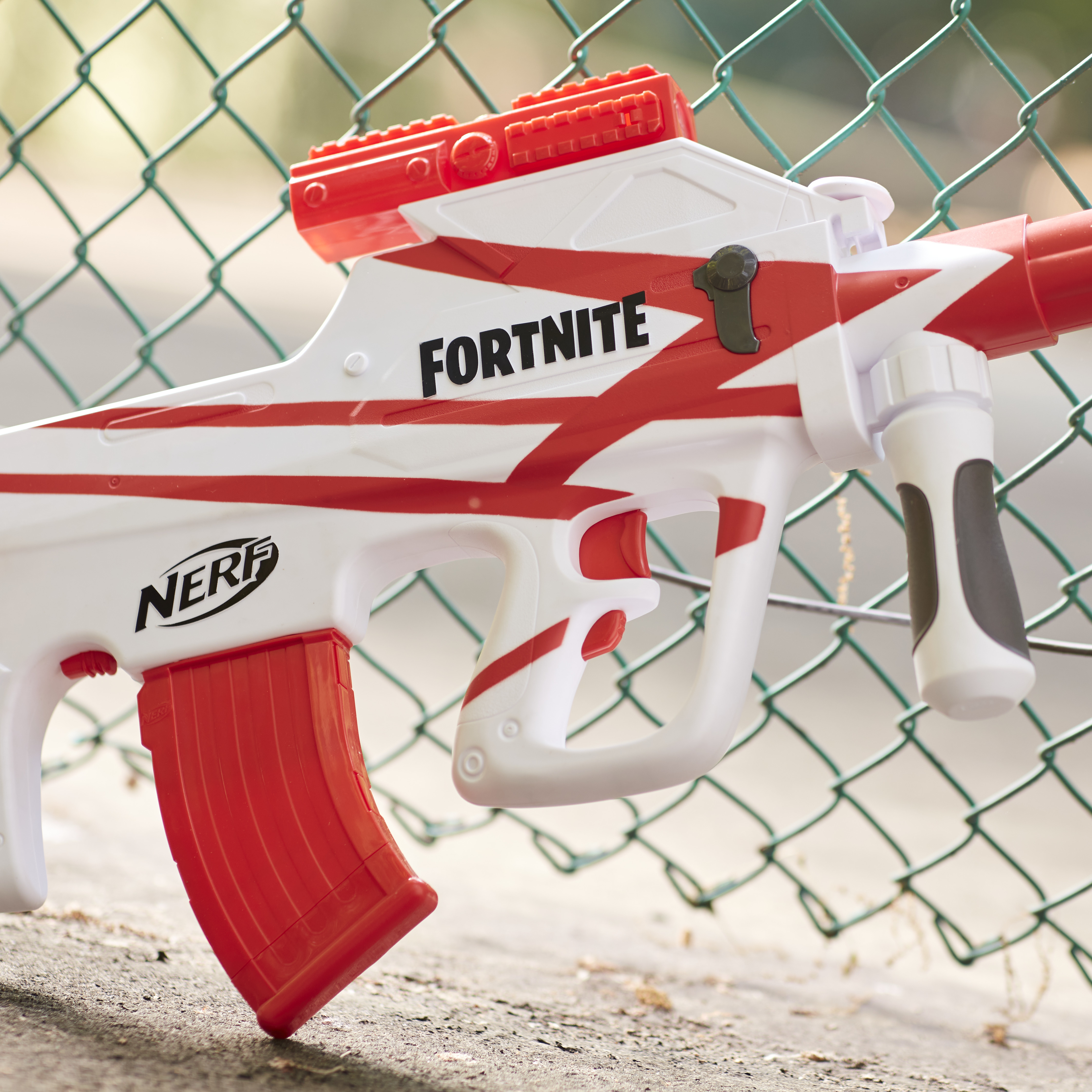 Biało-czerwony pistolet Nerf Fortnite opiera się o ogrodzenie z siatki drucianej. Widoczne jest słowo 'FORTNITE'.