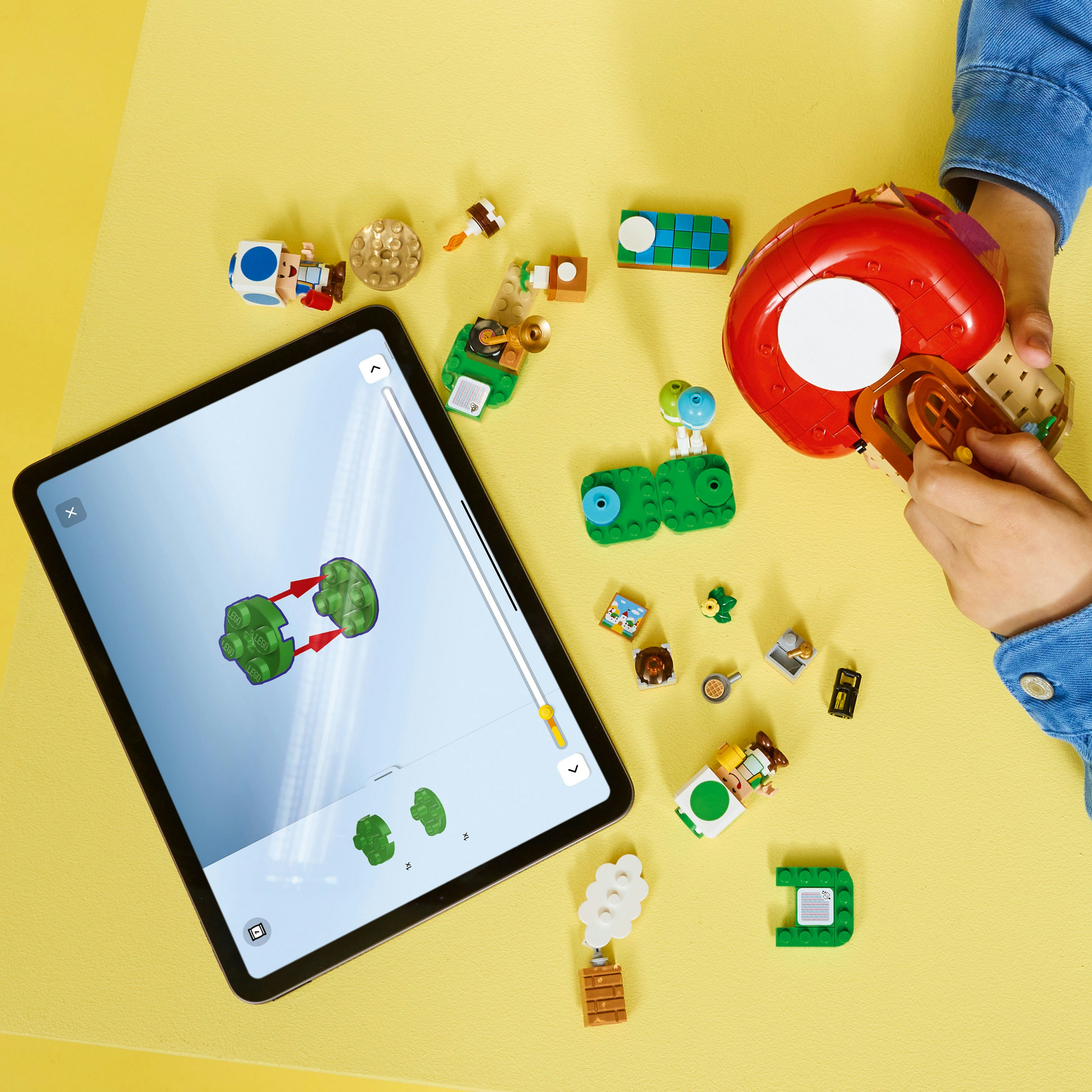 Ein Tablet zeigt Bauanleitungen. Lego-Teile umgeben es. Die Hand eines Kindes hält ein rotes, pilzförmiges Teil.