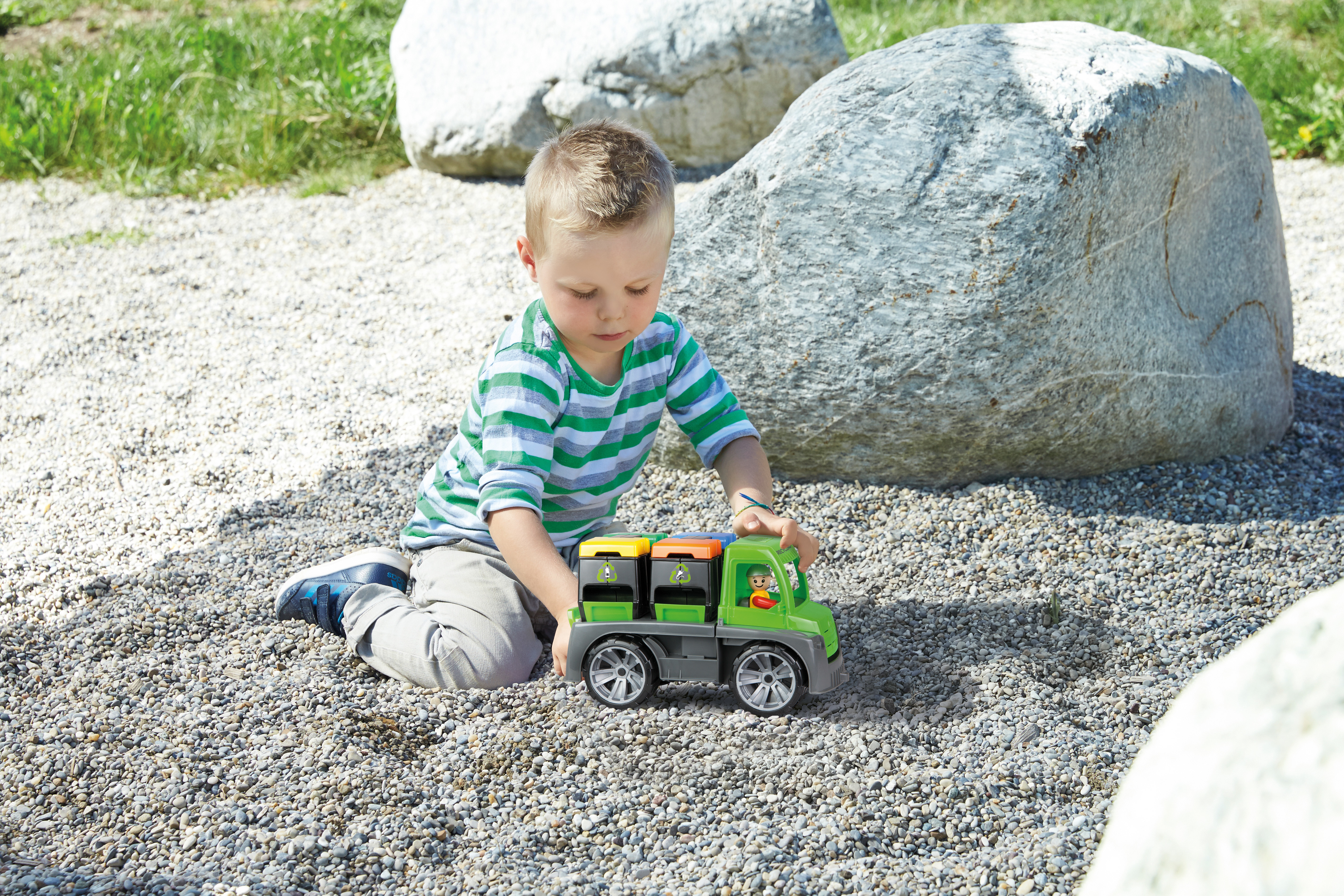 Chłopiec w zielono-pasiastej koszuli bawi się zieloną zabawką ciężarówką na kamykach, w pobliżu dużych skał i trawy.