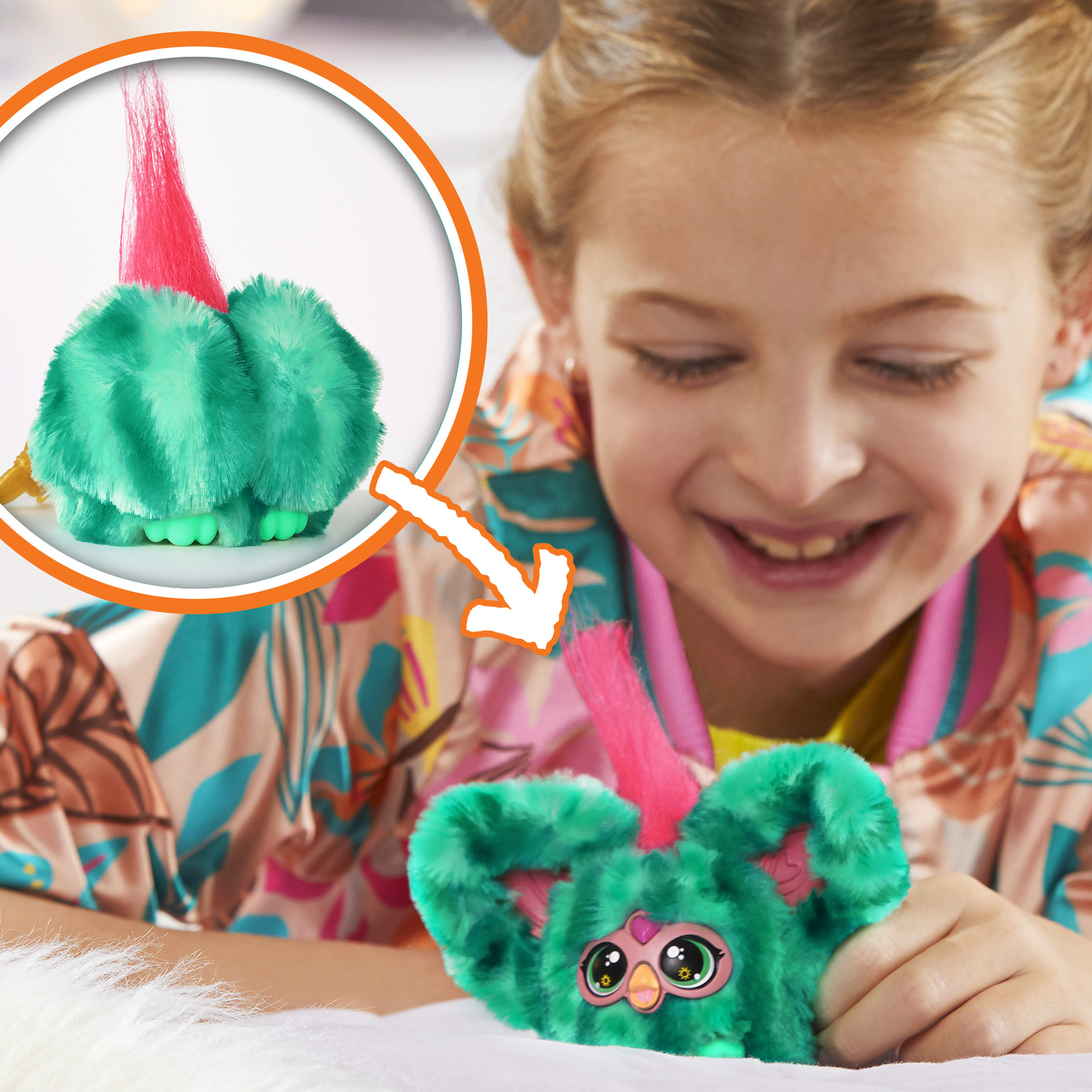 Una niña sonríe a un peluche verde con pelo rosa. El juguete tiene ojos grandes y está sobre una superficie blanca.