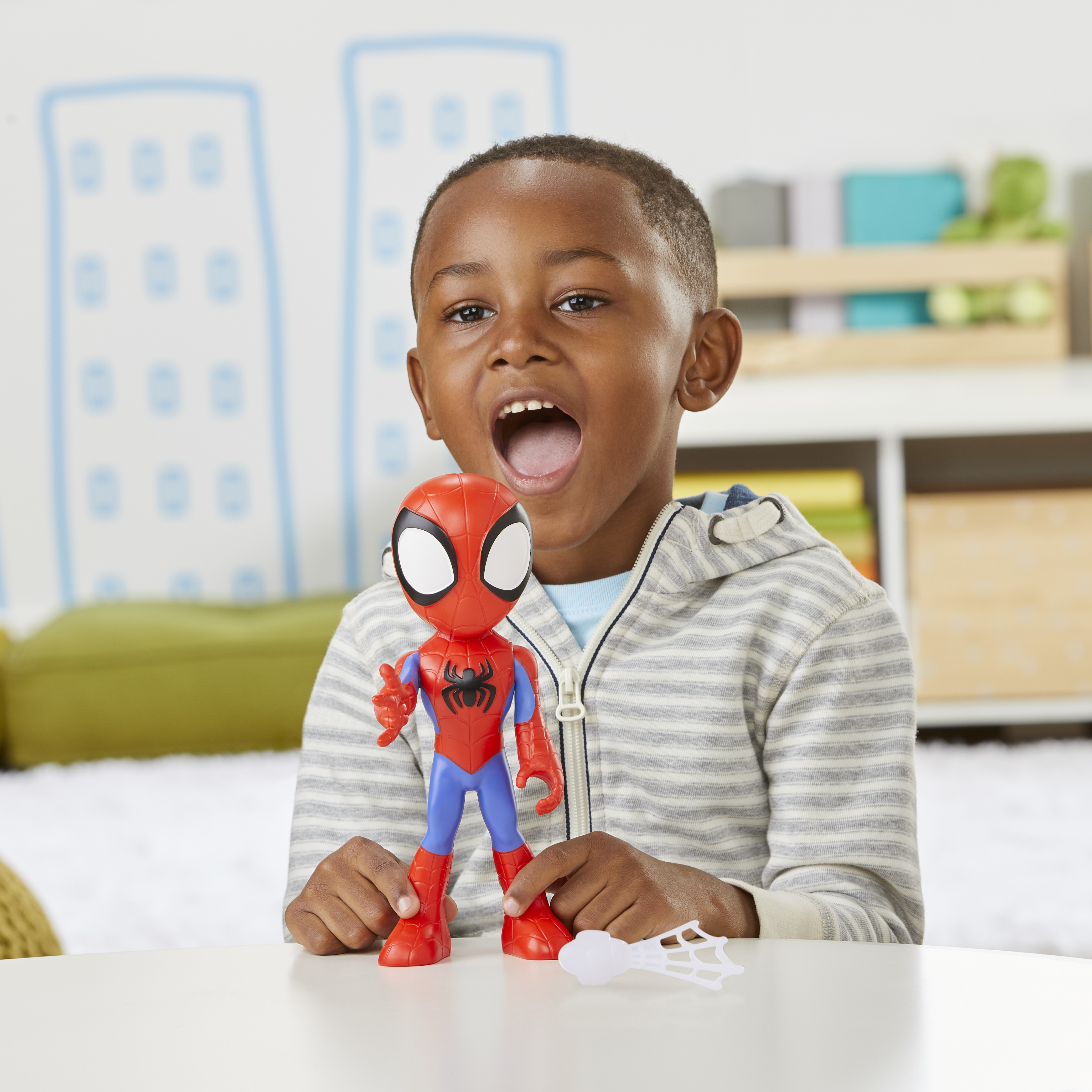 Un niño sostiene un juguete de Spider-Man, con la boca abierta. El juguete y el niño están en una mesa. El fondo incluye dibujos de edificios.