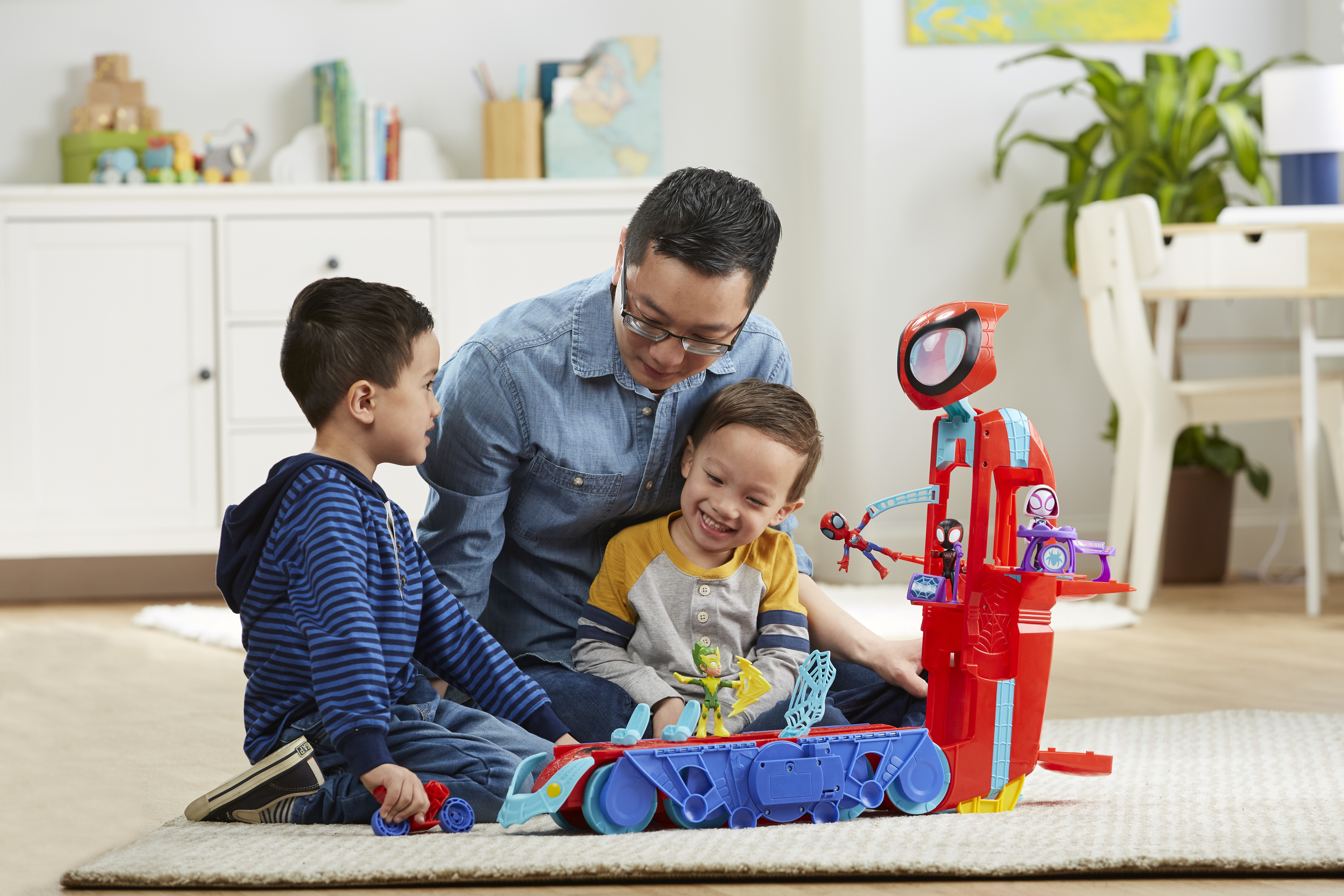 Mężczyzna i dwoje dzieci bawią się zabawką Spider-Mana. Zabawka jest czerwona, niebieska. Tło przedstawia białą szafkę.