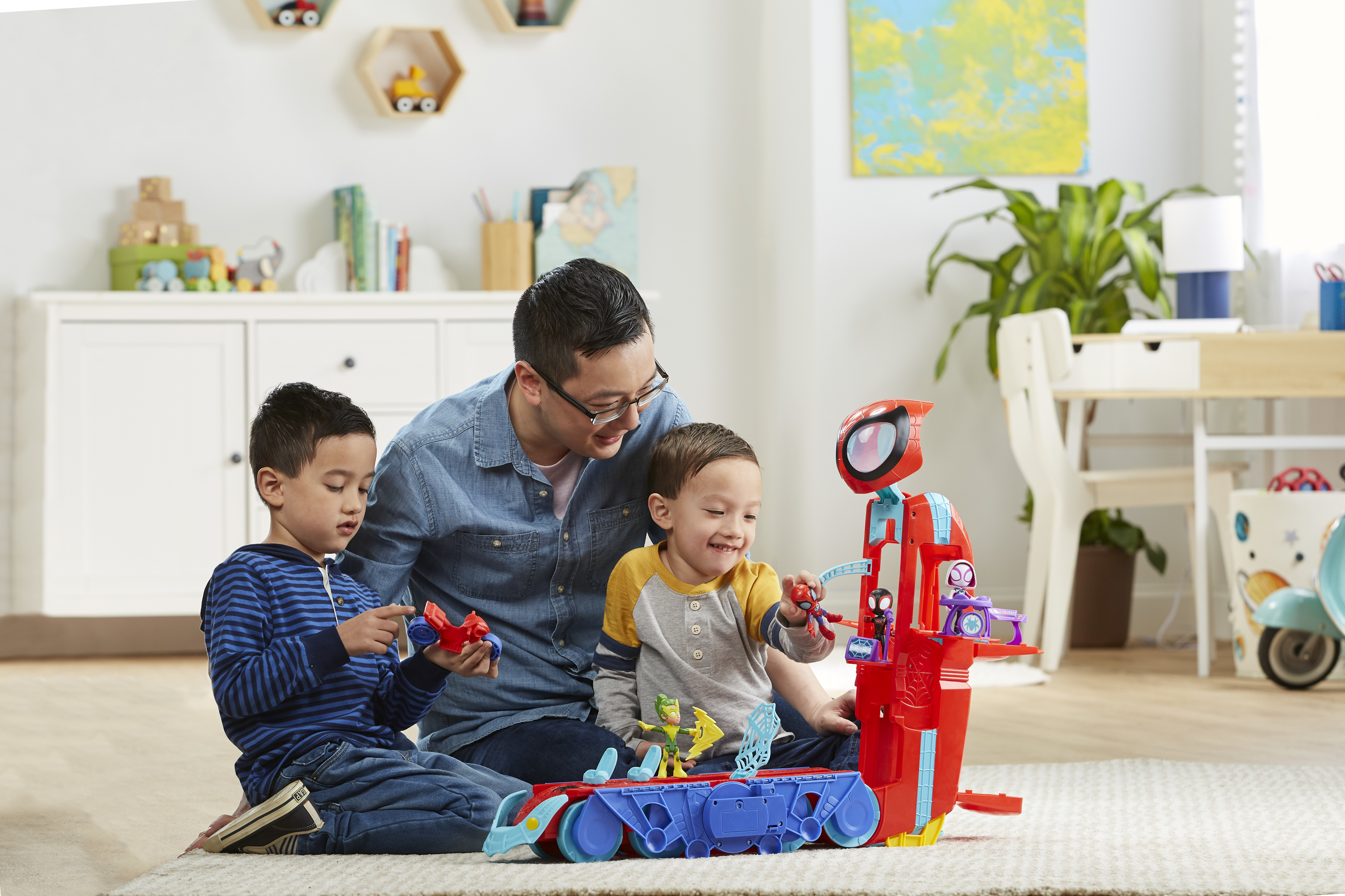 Mężczyzna i dwóch chłopców bawią się zabawkami Spidermana. Czerwony zestaw zabawek jest na pierwszym planie. Biała szafka w tle.