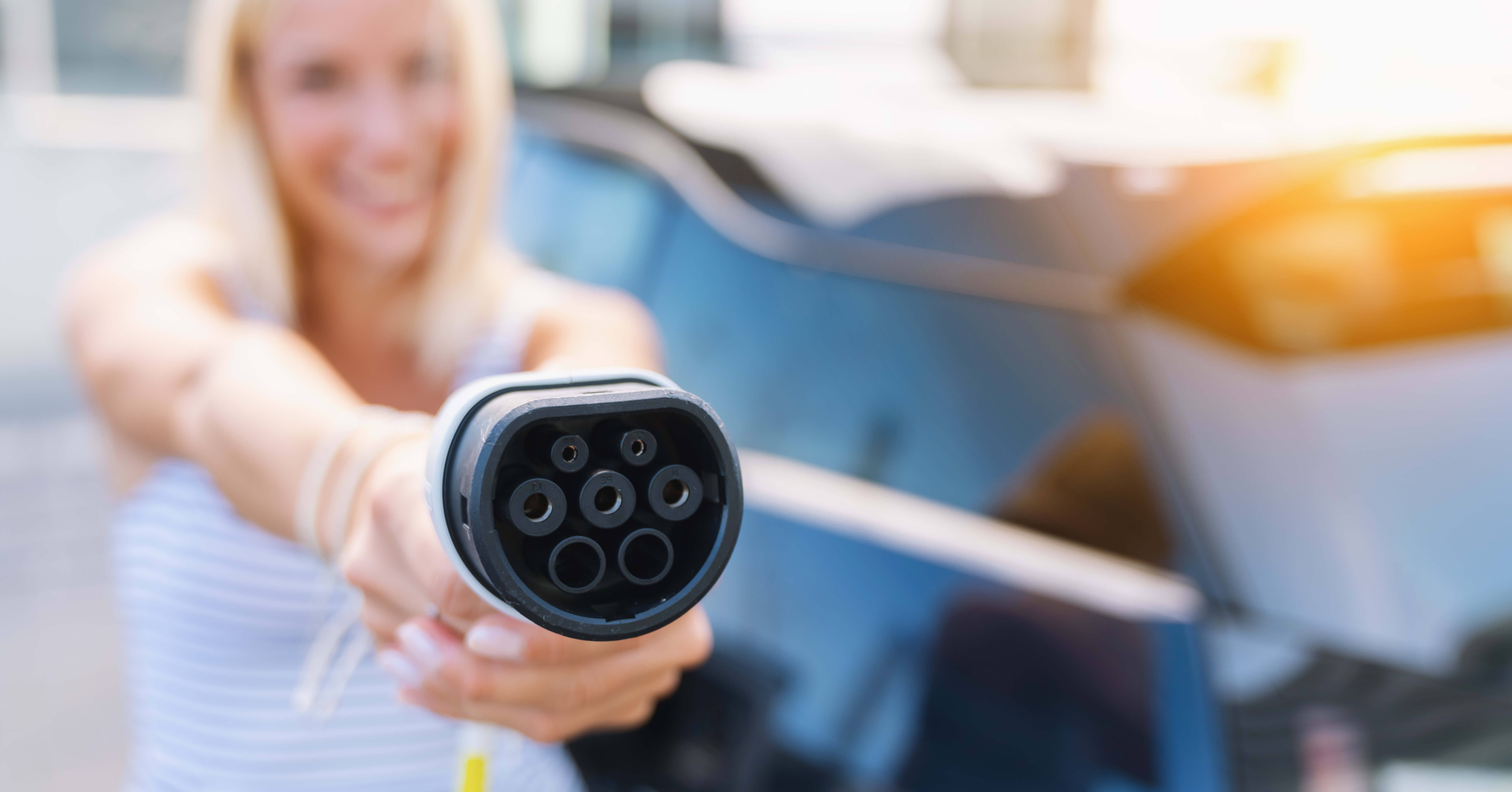 Una donna tiene un connettore di ricarica per veicoli elettrici verso la fotocamera. Un'auto blu sullo sfondo.