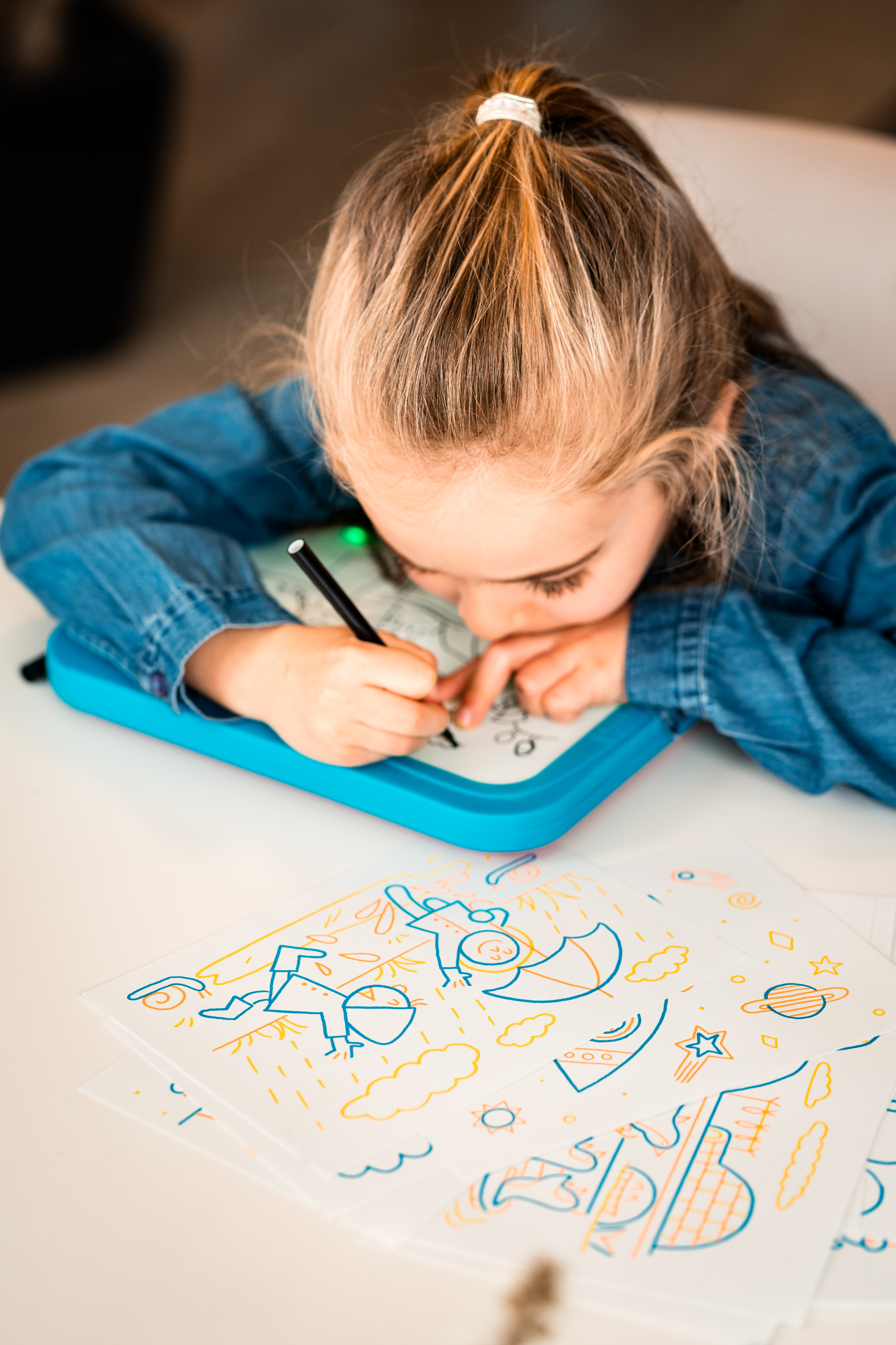 Dziewczynka rysuje na tablecie. Rysunki są na papierze. Dziewczynka ma na sobie niebieską koszulę i blond włosy.