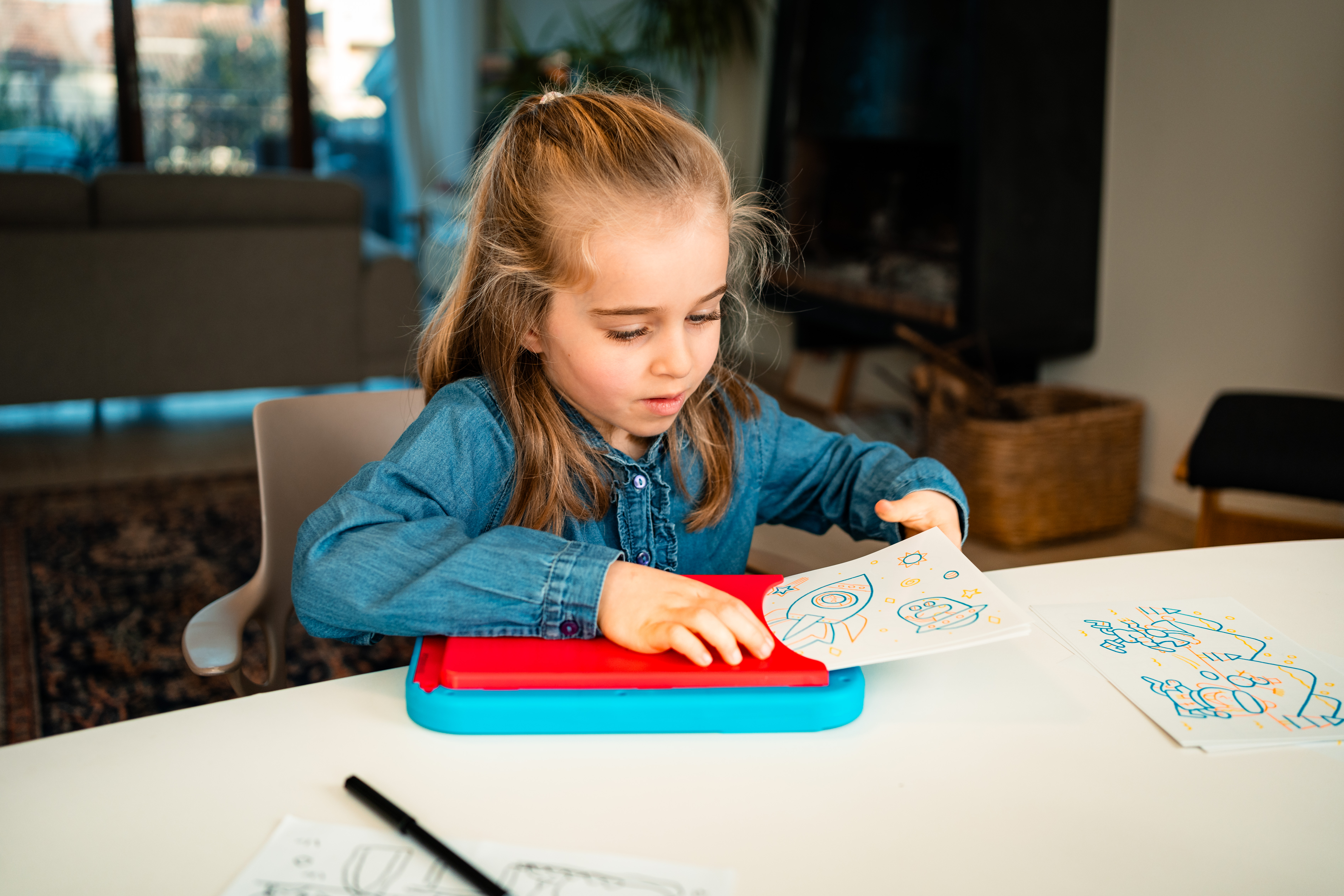 Dziewczyna rysuje rakietę. Jest w pokoju ze stołem, krzesłem i rysunkiem statku kosmicznego.