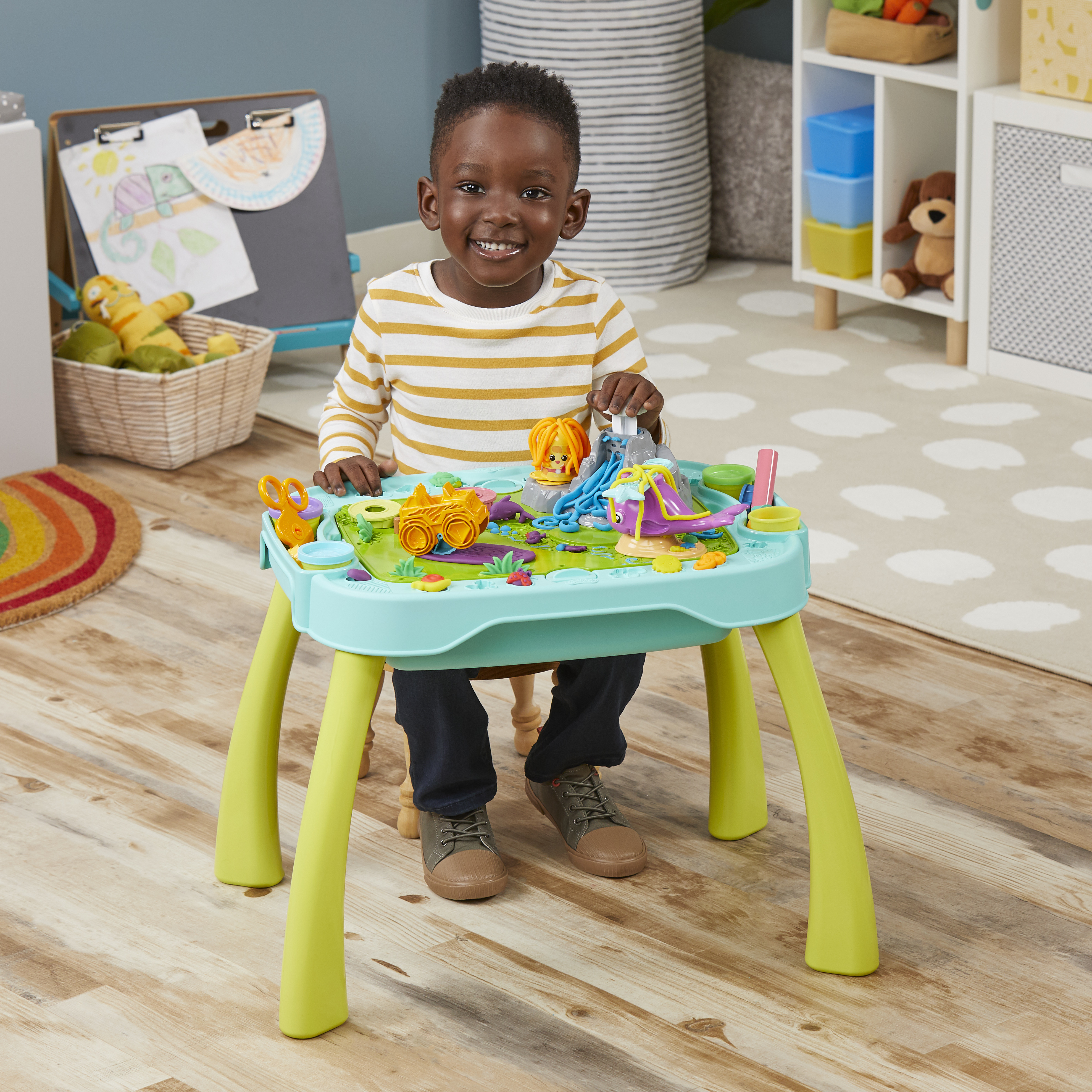 Un niño sonriente está sentado a una mesa de juegos. La mesa tiene juguetes. Una pizarra y una cesta de juguetes están al fondo.